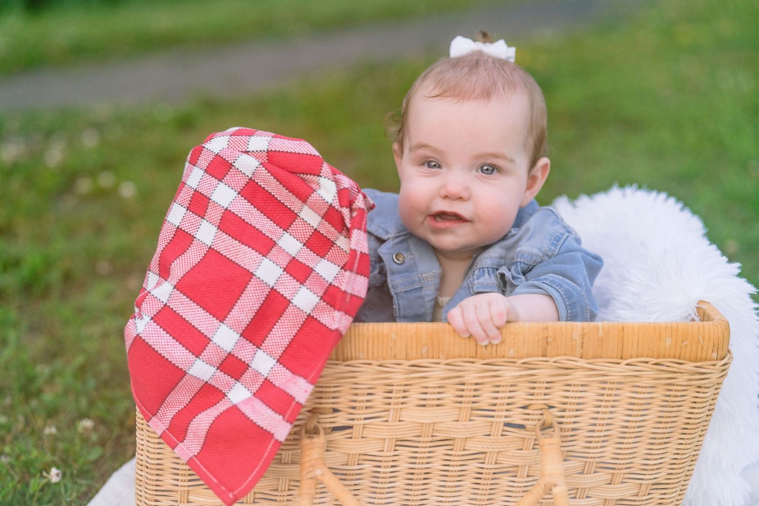 Baby in denim jacket leans against wicker picnic basket on blanket outdoors.