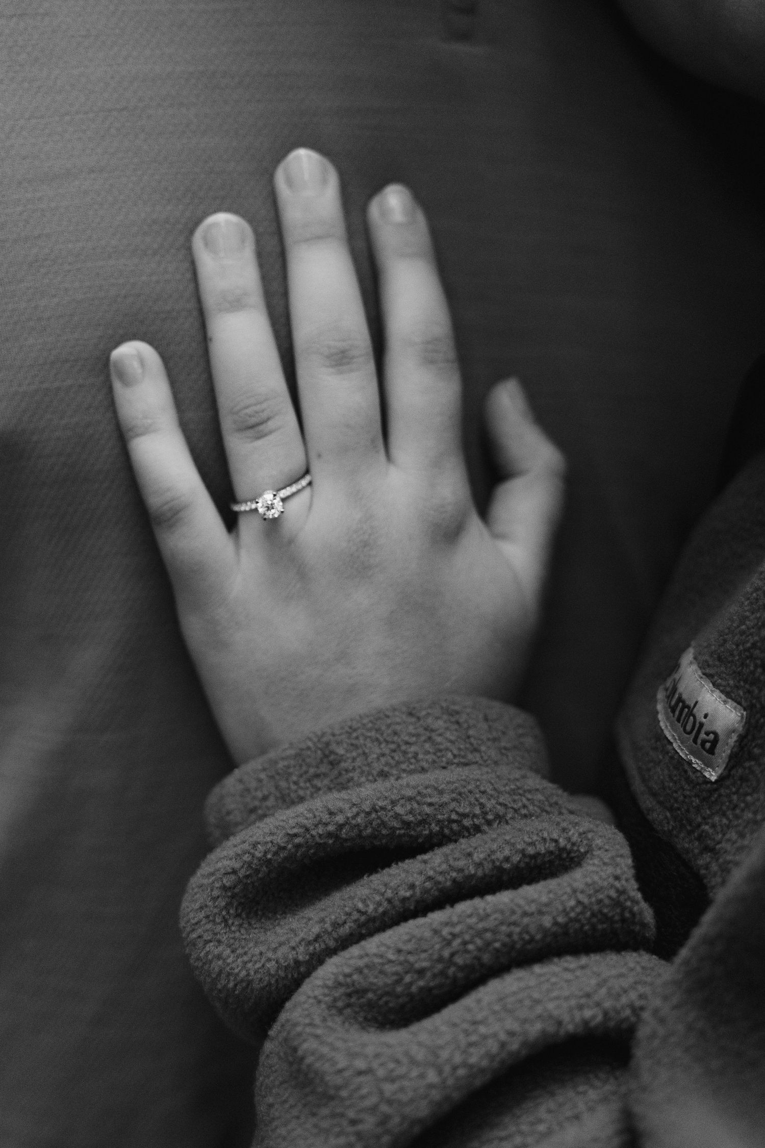 A close-up black and white photo of a hand wearing an engagement ring against a dark background.