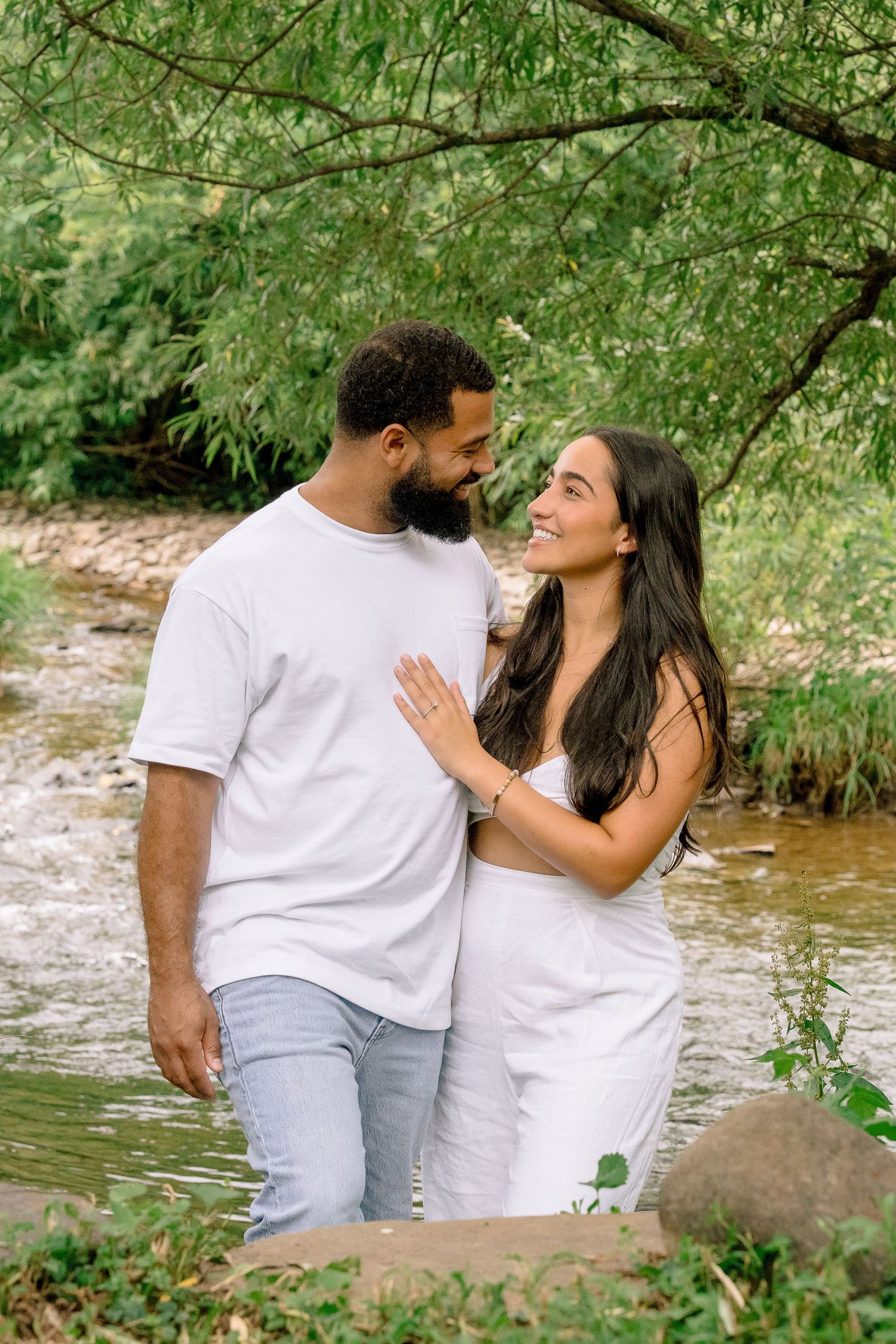 A couple shares a romantic moment by a stream in nature, both wearing white outfits against lush green surroundings.