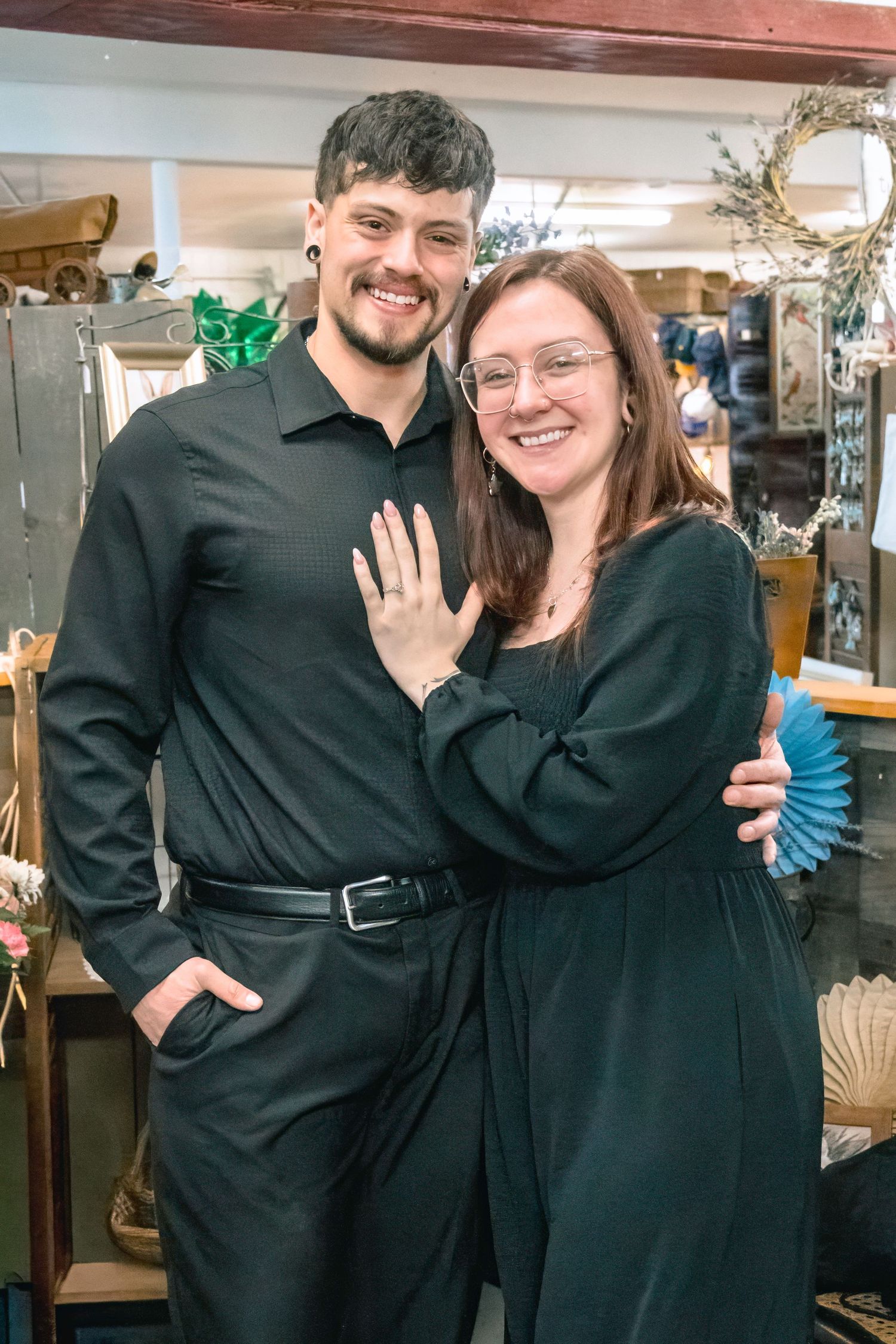 Happy couple in matching black outfits smiling in a shop, woman showing off engagement ring on her hand.