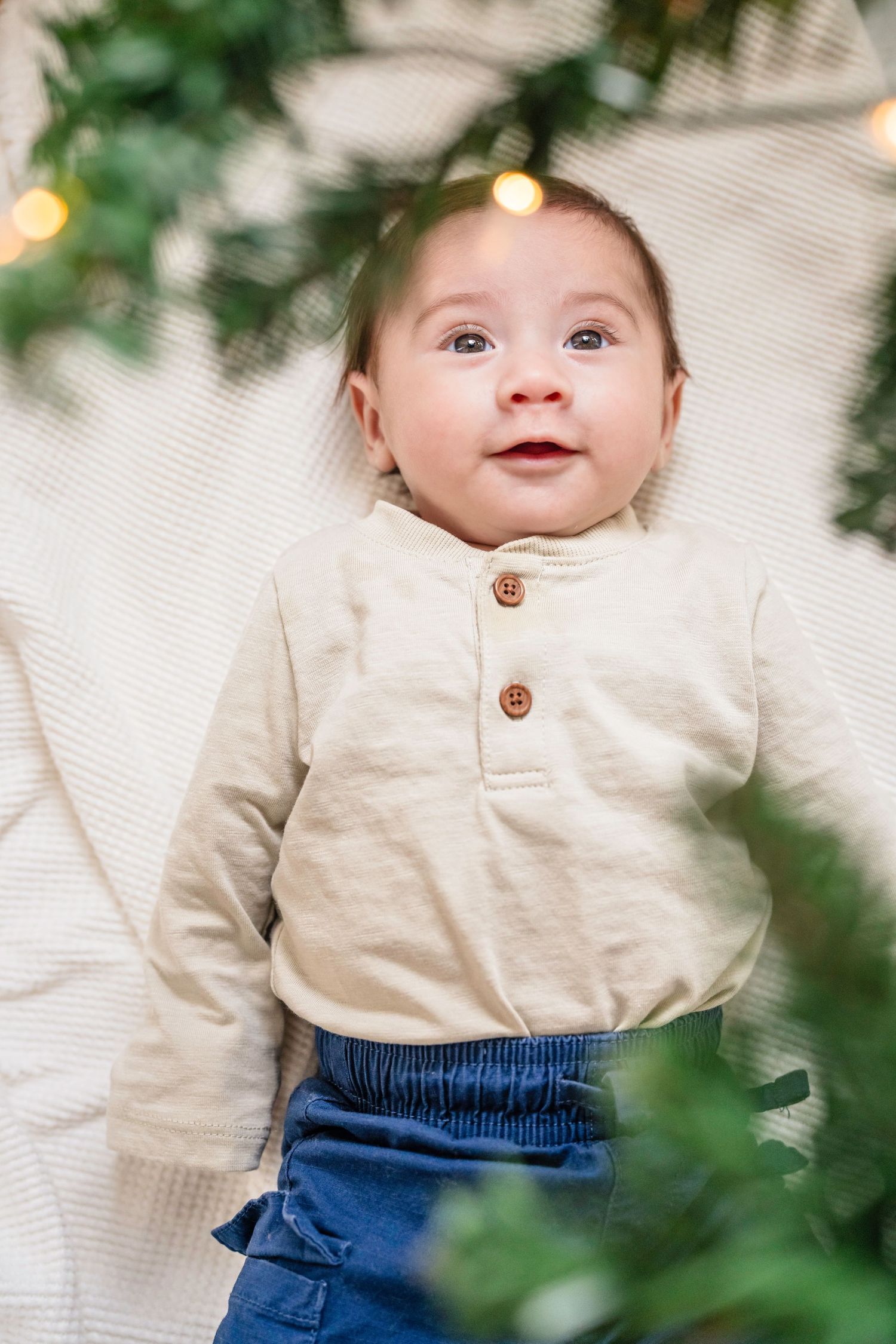 Baby wearing cream henley shirt and navy pants smiles while lying on white blanket with greenery and lights in background. Christmas.