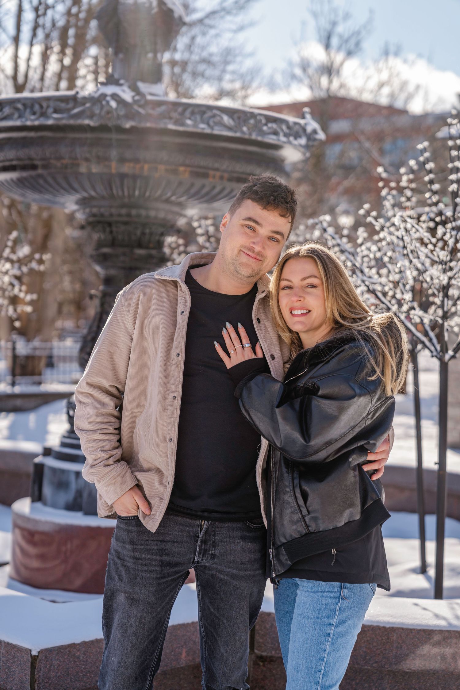Happy couple posing in front of a snowy fountain outdoors in winter, wearing casual jackets and jeans.