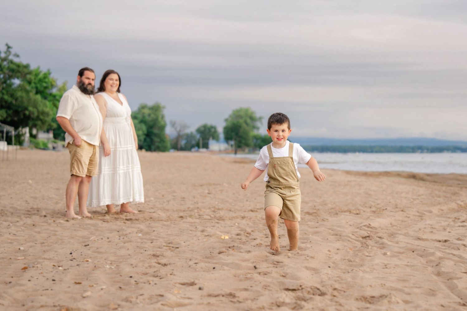 A small child runs joyfully on a sandy beach at dusk while two figures stand in the background near the water.