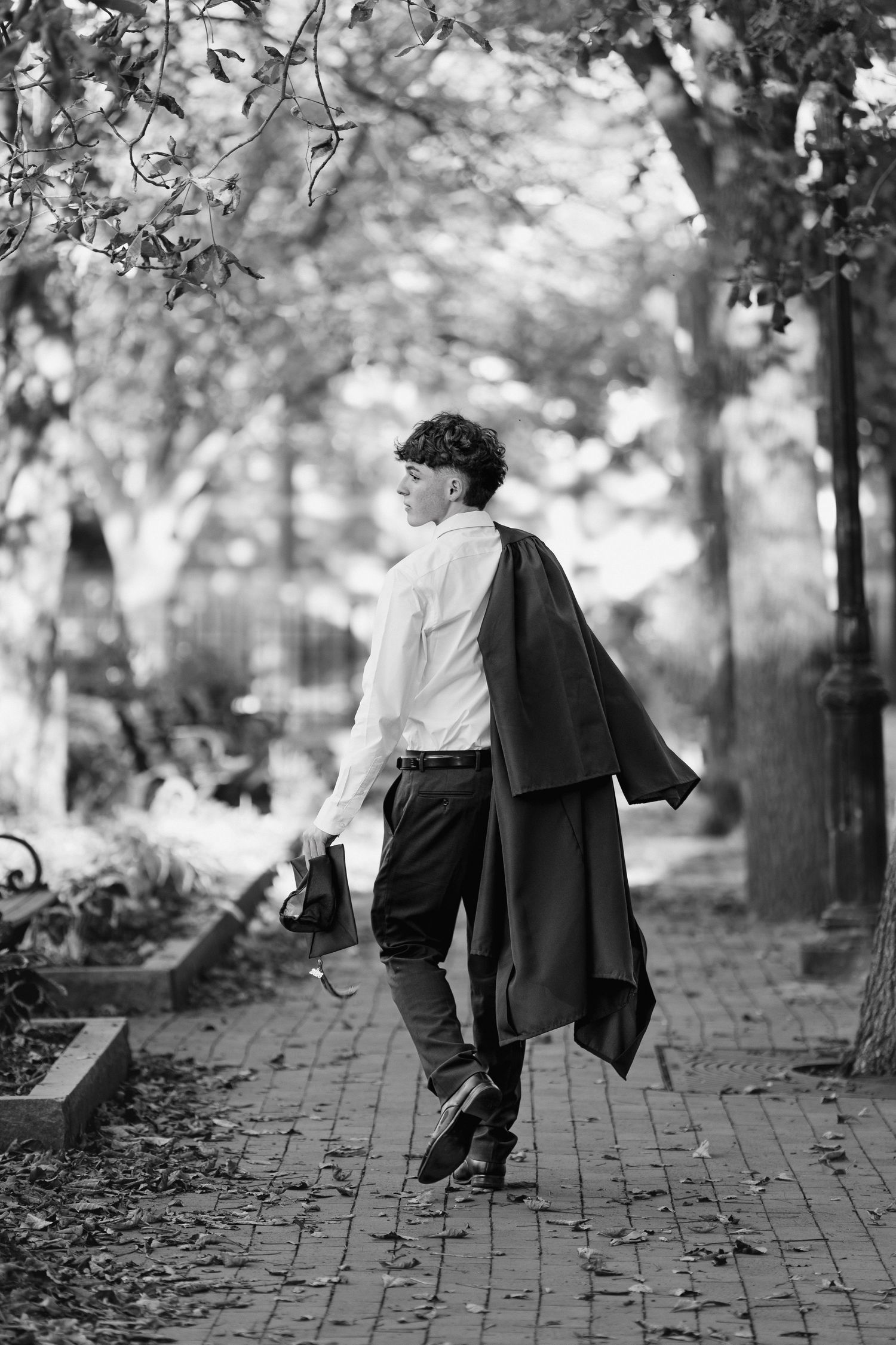 Black and white photo of someone walking on a brick path through a park, wearing a draped coat over their shoulder.