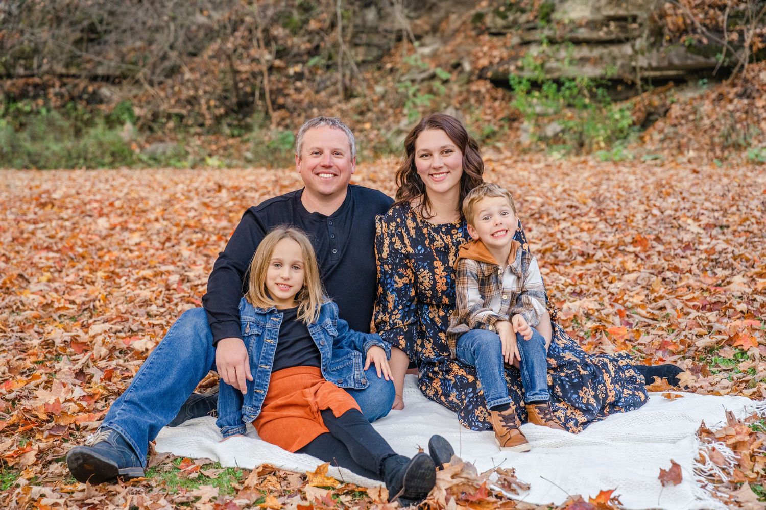 A family sits together on a white blanket surrounded by colorful autumn leaves in an outdoor setting.