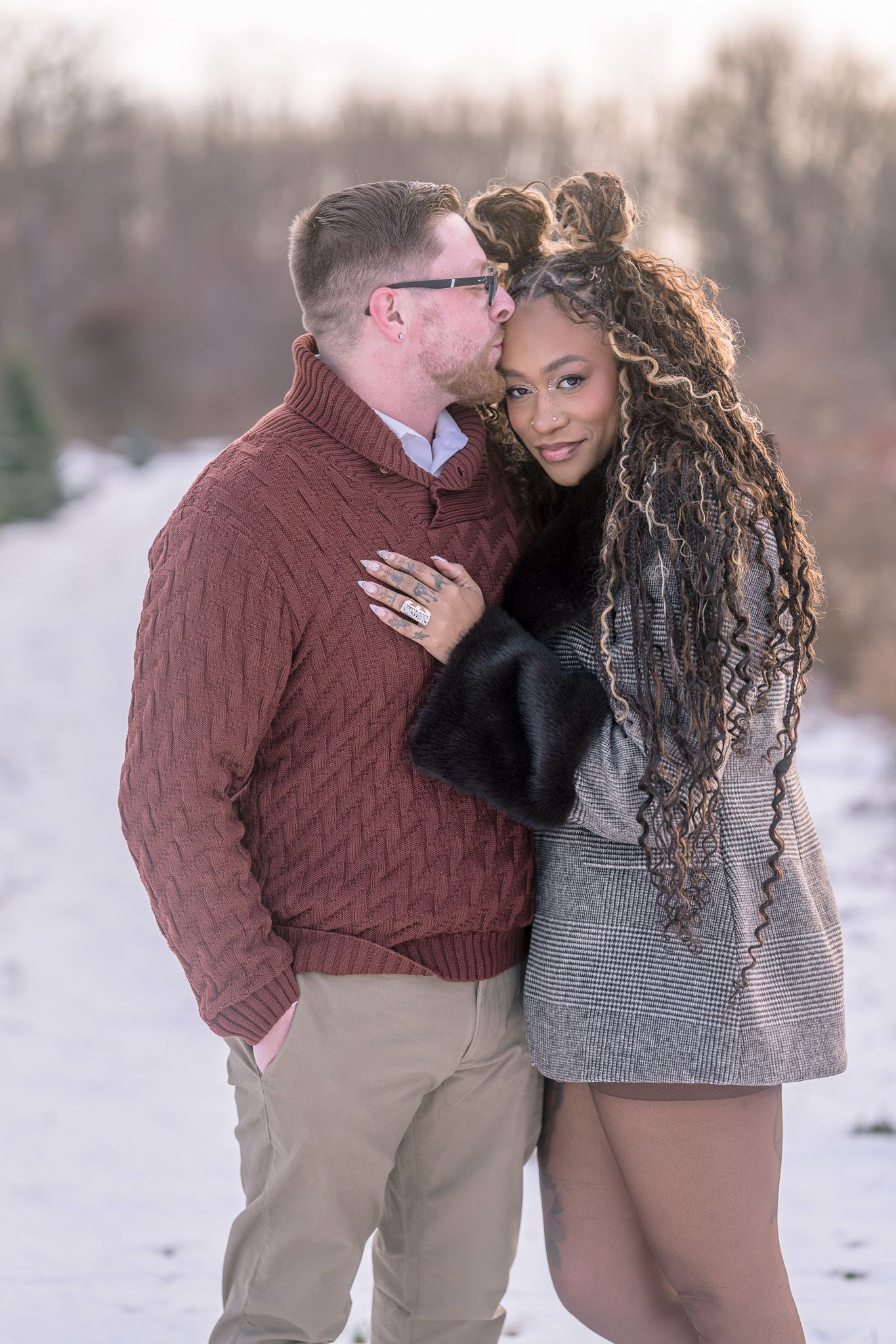 A couple embraces in a romantic winter photoshoot wearing burgundy and grey outfits against a snowy backdrop.
