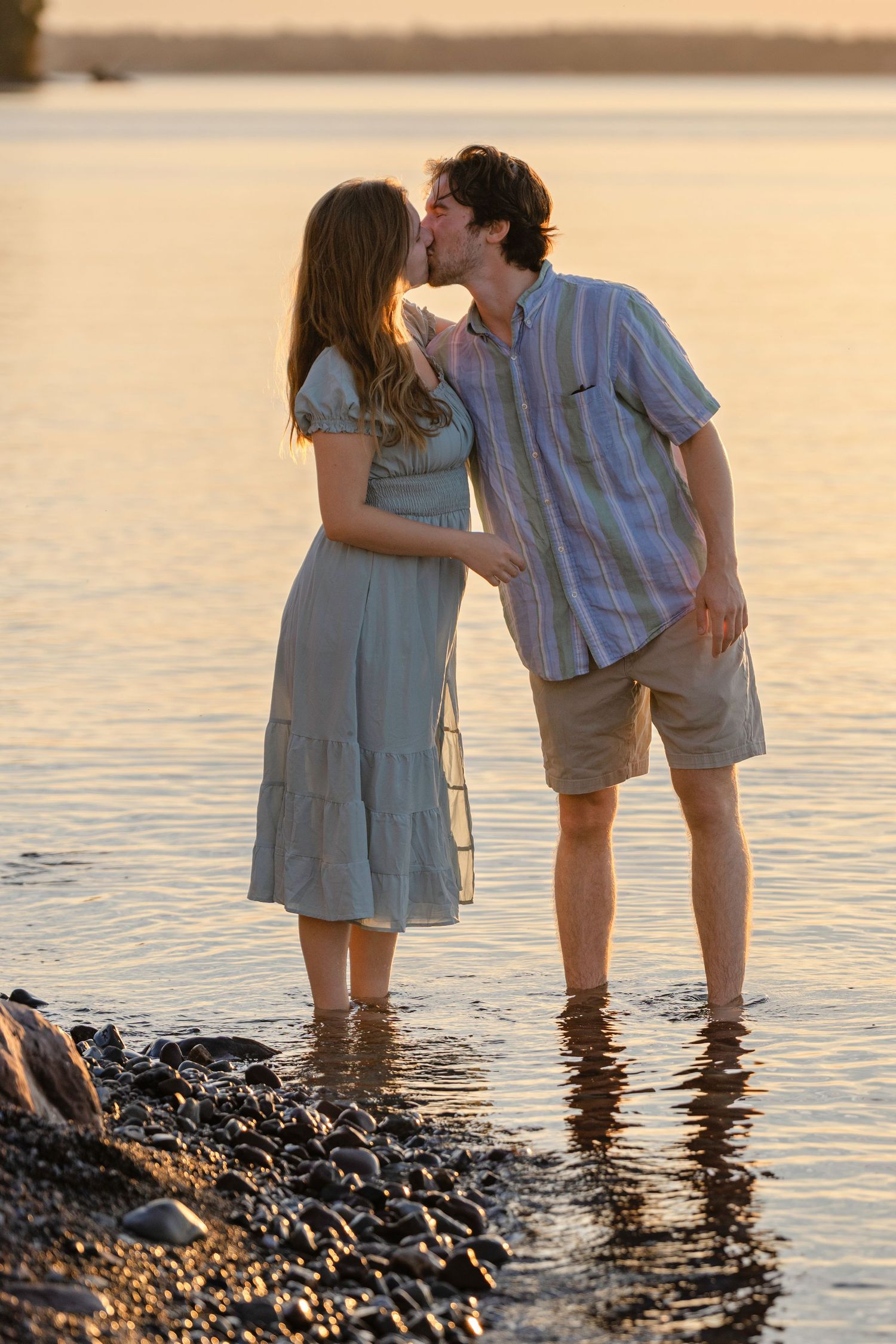Romantic couple standing in shallow water at sunset, with her in a sundress and him in casual summer attire.