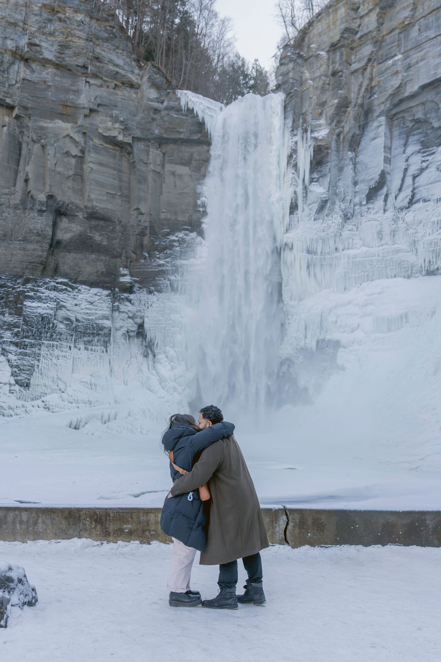 A person kneels in the snow in front of a frozen waterfall during a winter proposal moment.