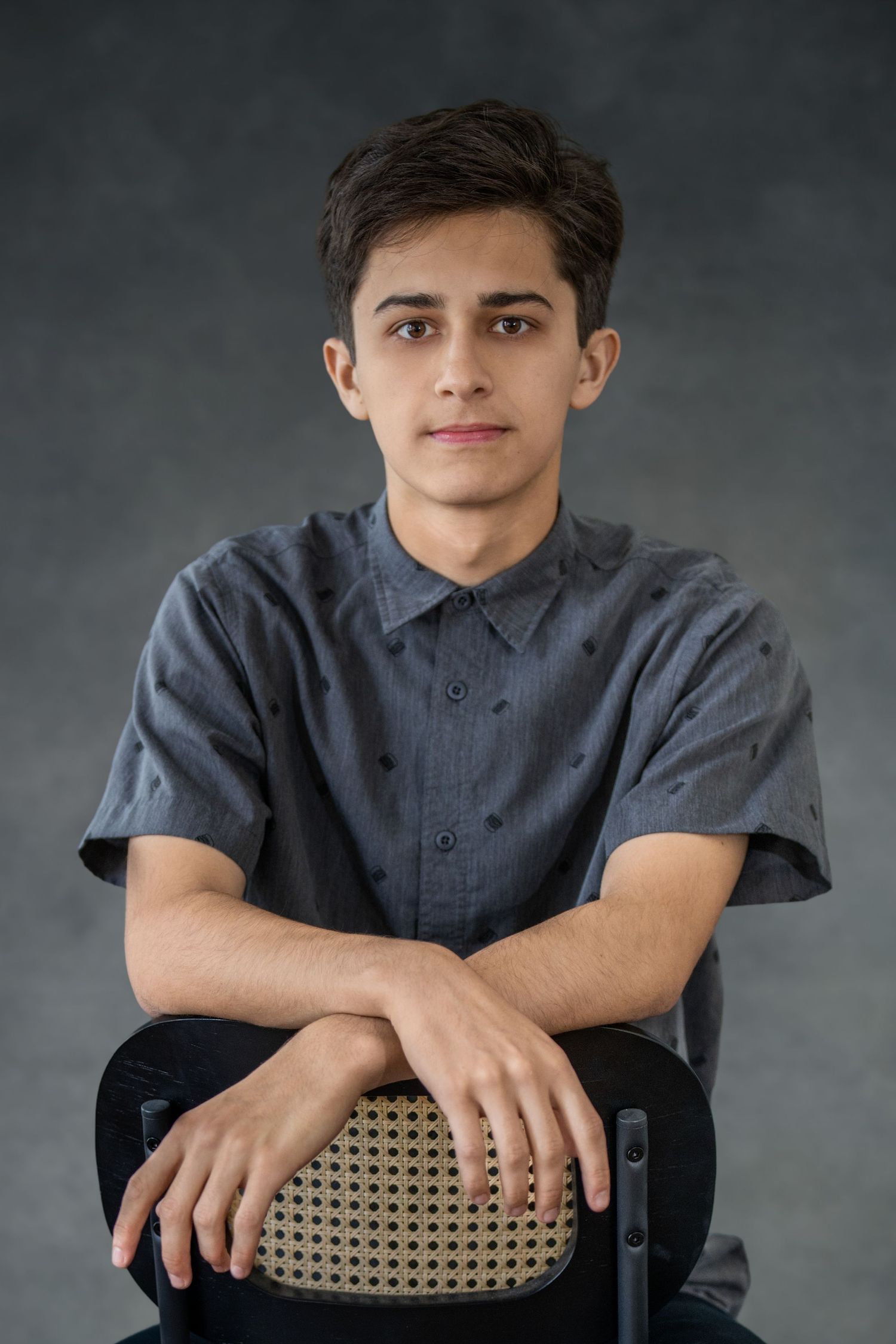 Young musician in a gray button-up shirt poses against dark background while seated on a chair.
