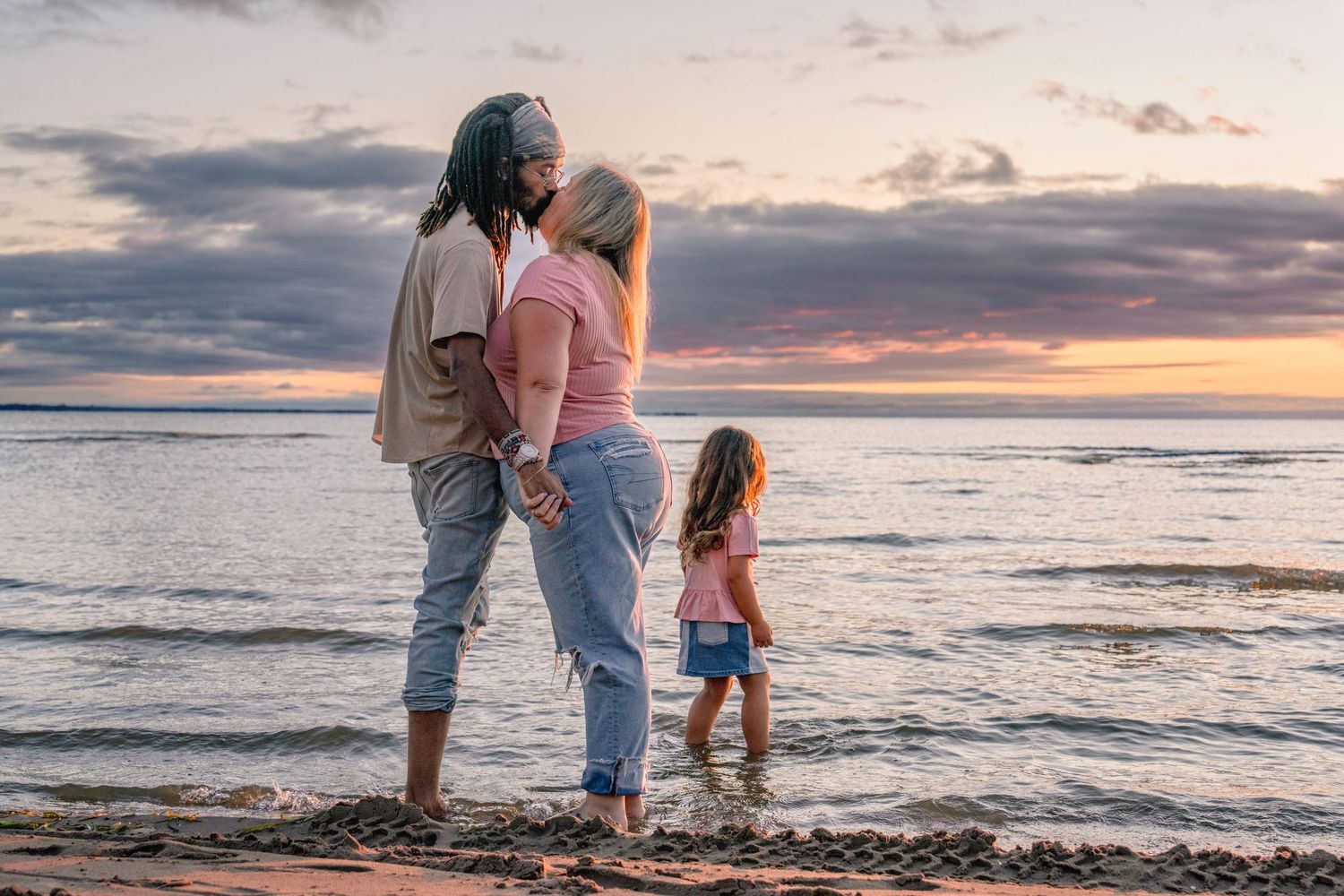 Family members walk hand in hand along beach shore during colorful sunset.