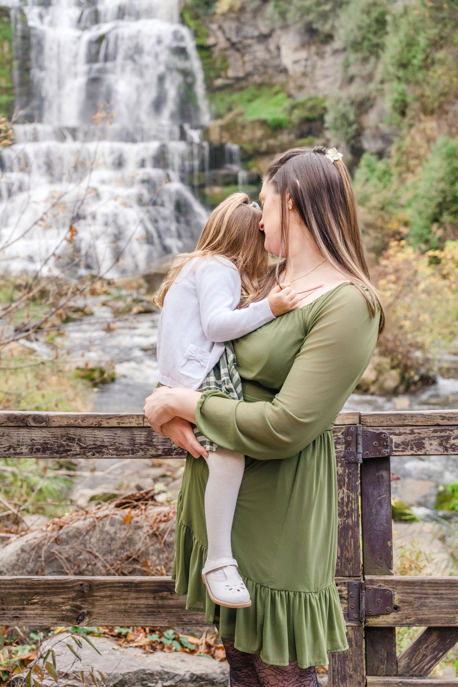 A mother in an olive green dress holds her baby near a cascading waterfall in a scenic outdoor setting.