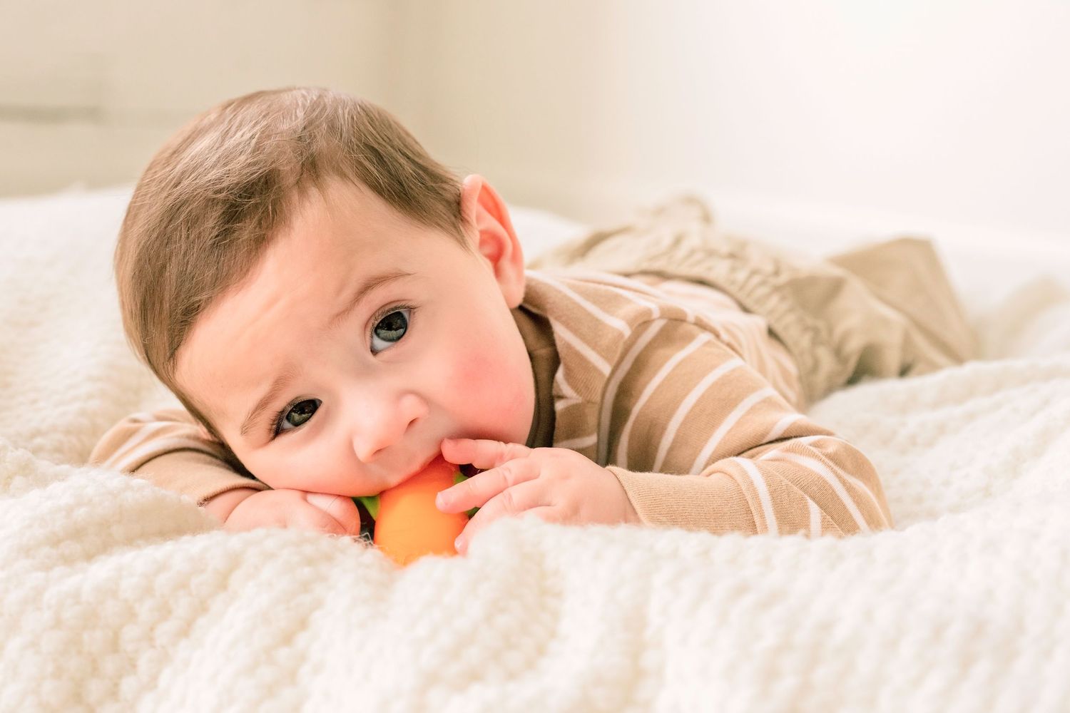 Cute baby in striped onesie lying on white blanket chewing orange toy.