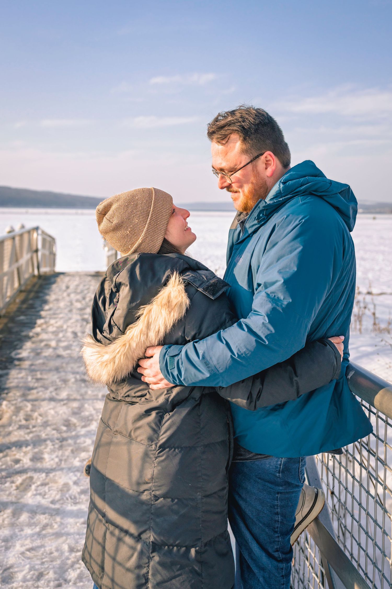 Romantic couple embracing on snowy pier in winter, smiling at each other under blue sky.