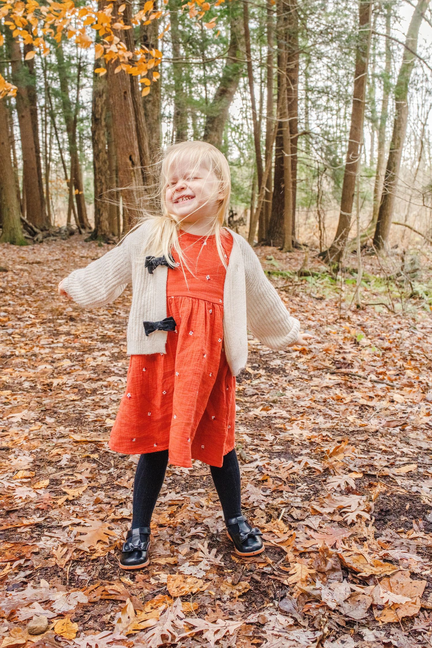 A child wearing a bright orange dress and boots plays in fallen autumn leaves on a forest floor.