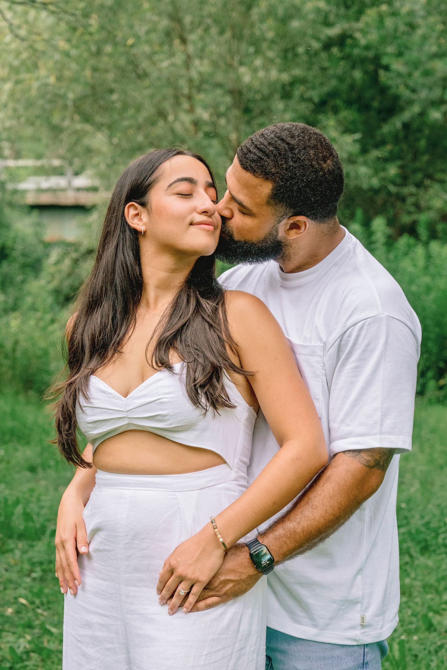 A couple wearing white t-shirts embrace lovingly in a lush green outdoor setting during a photo session.
