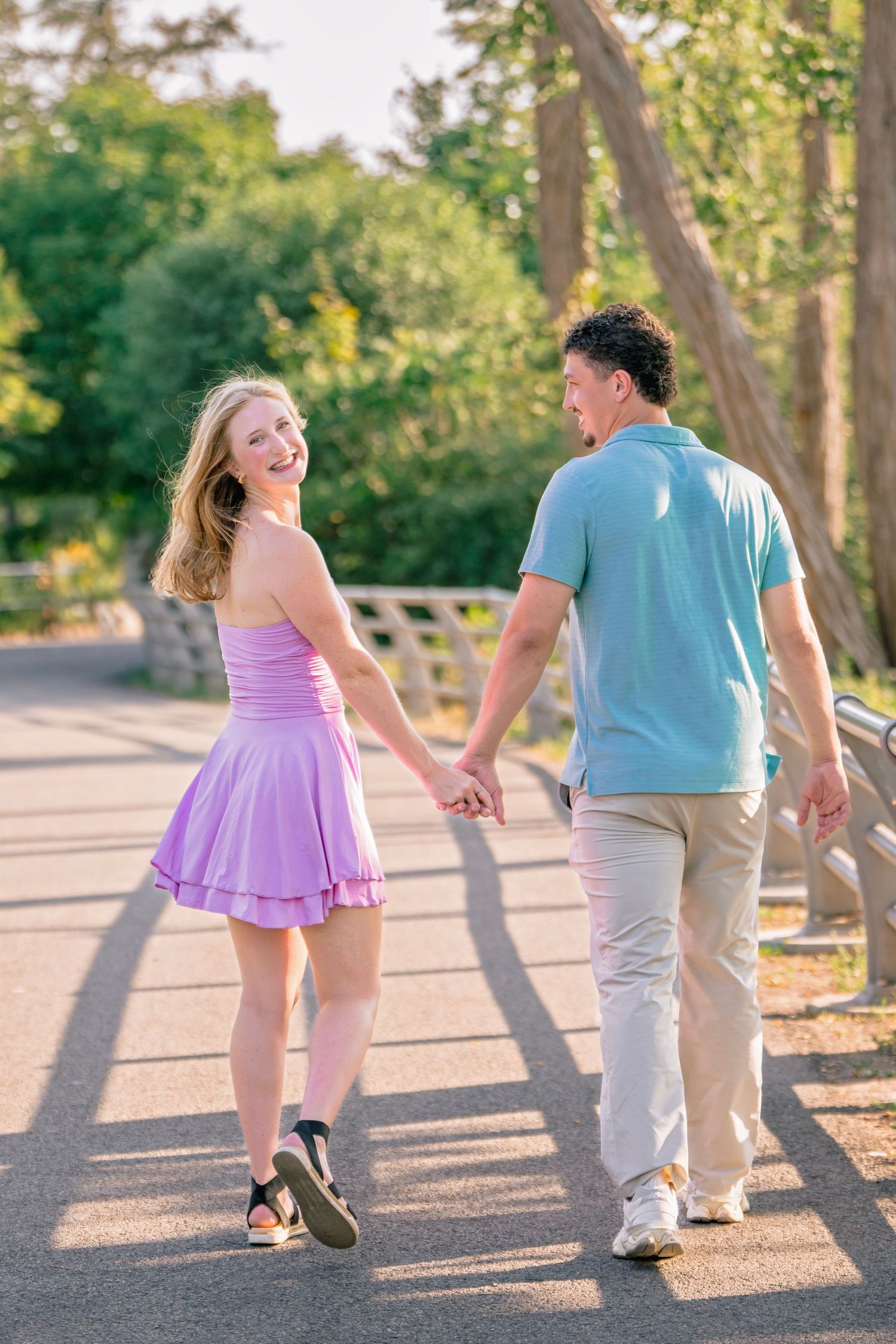 A couple walks hand in hand along a sunny path with trees casting shadows.