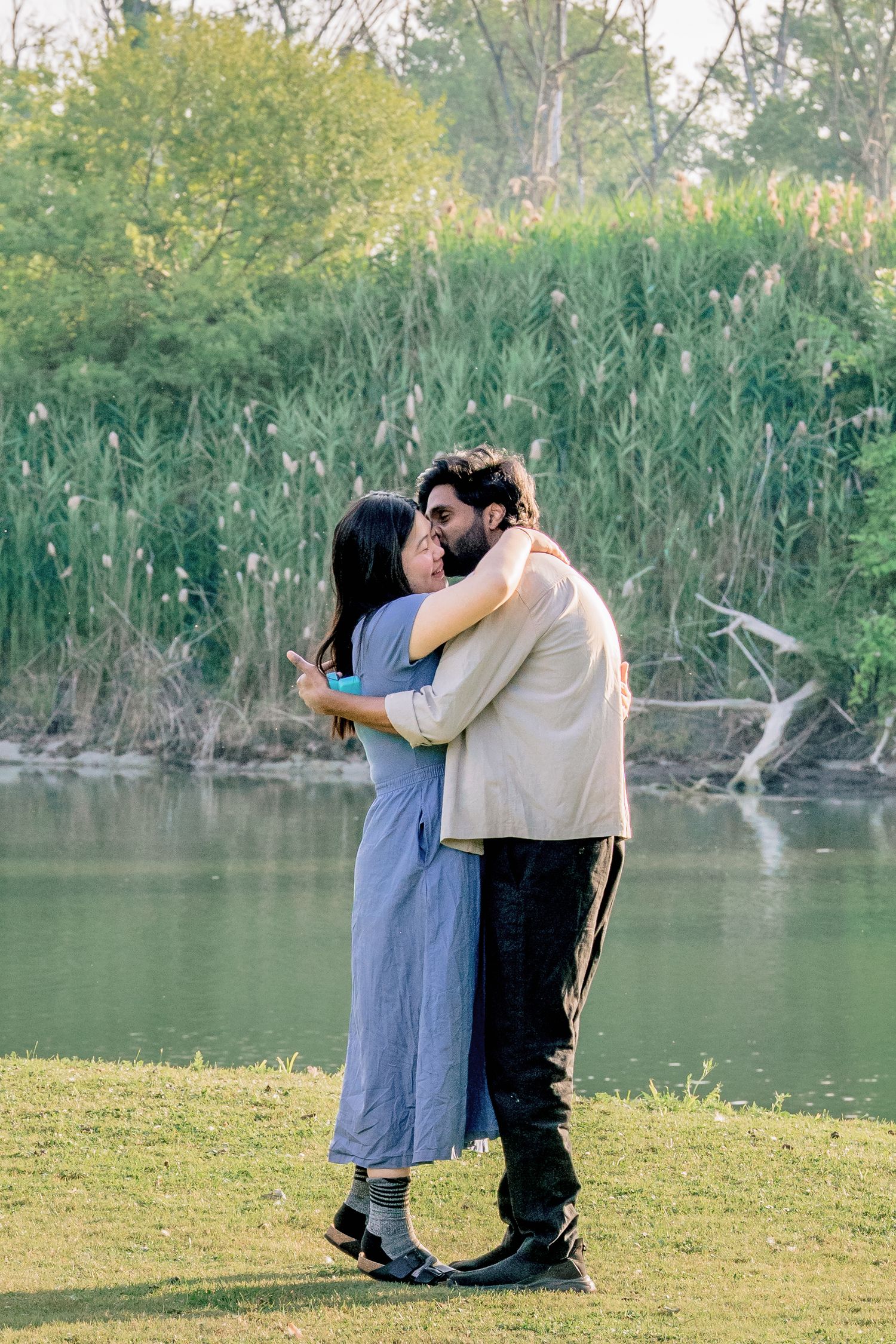 A couple embraces while standing by a tranquil lake surrounded by lush greenery and reeds.