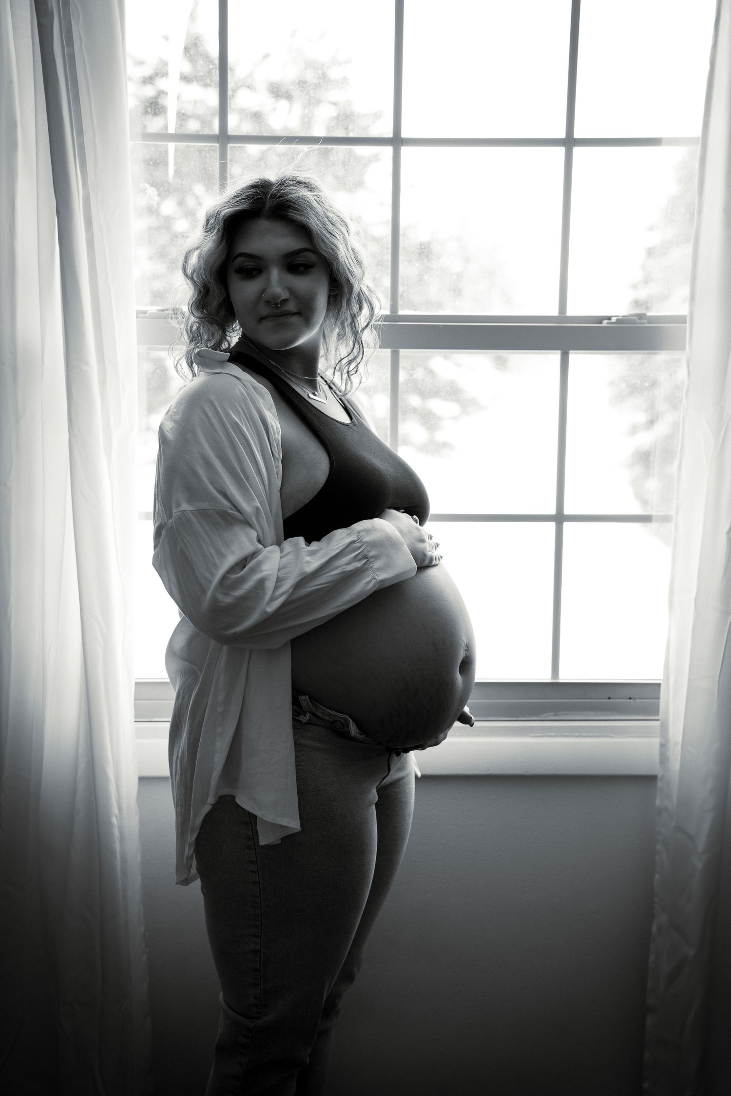 Black and white maternity portrait of an expectant mother standing by a bright window in casual attire.