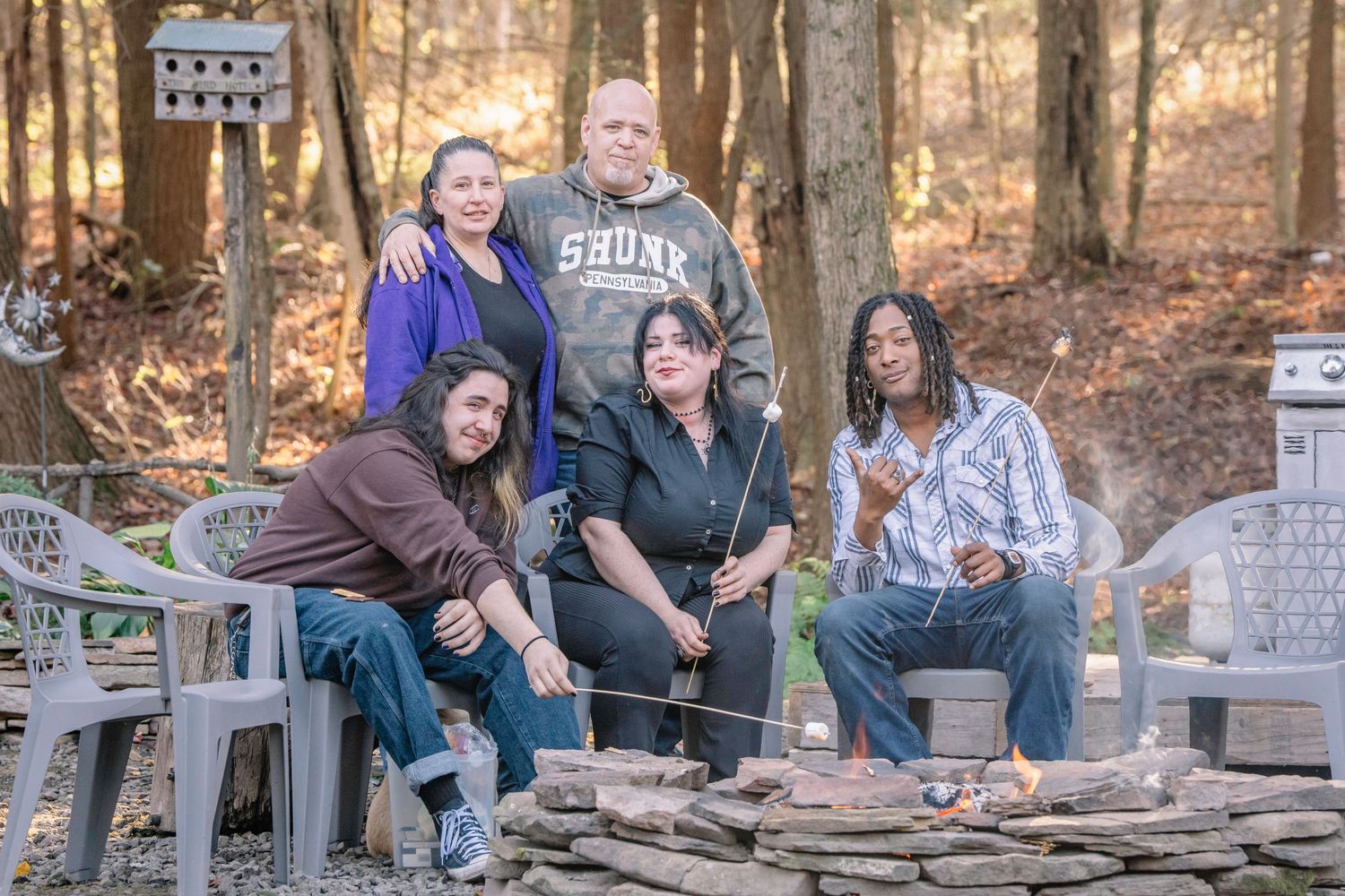 A group of people gather around picnic tables in a wooded outdoor setting during autumn.
