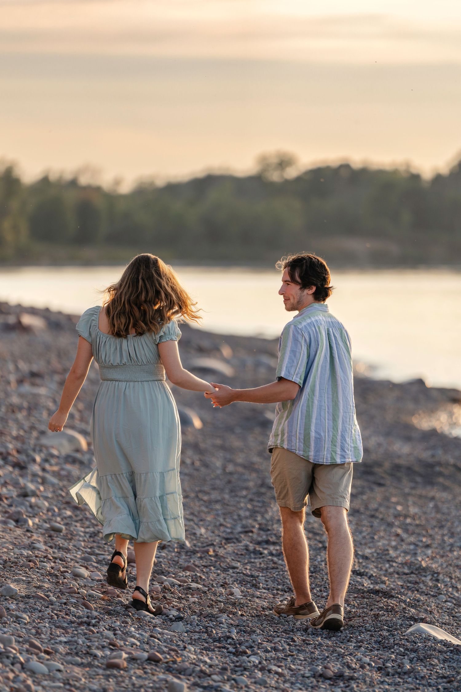 A couple walks hand in hand along a rocky beach shoreline at sunset wearing coordinating blue and khaki outfits.