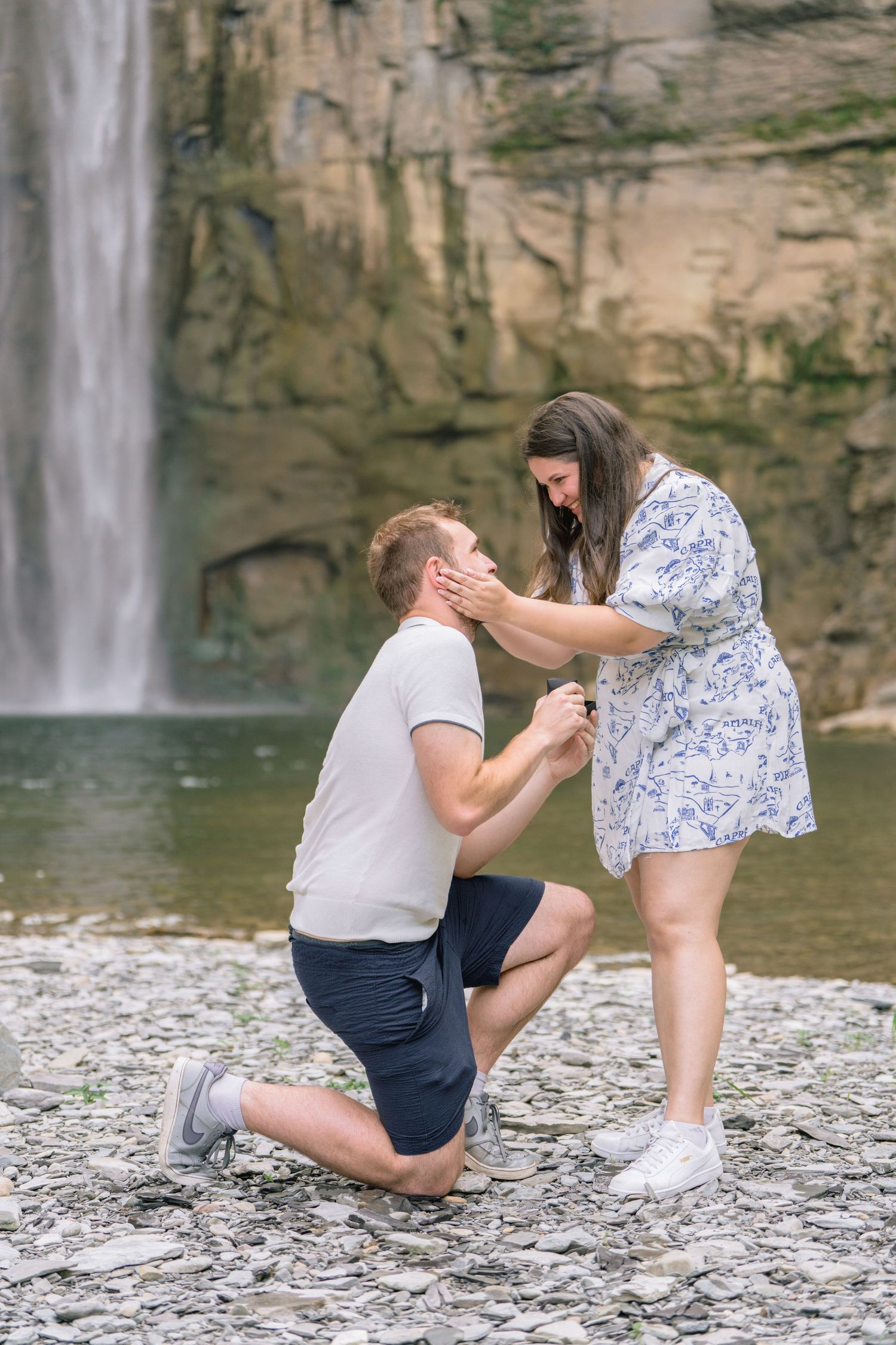 A romantic proposal sequence captured at a scenic waterfall location with rocky cliffs in the background.