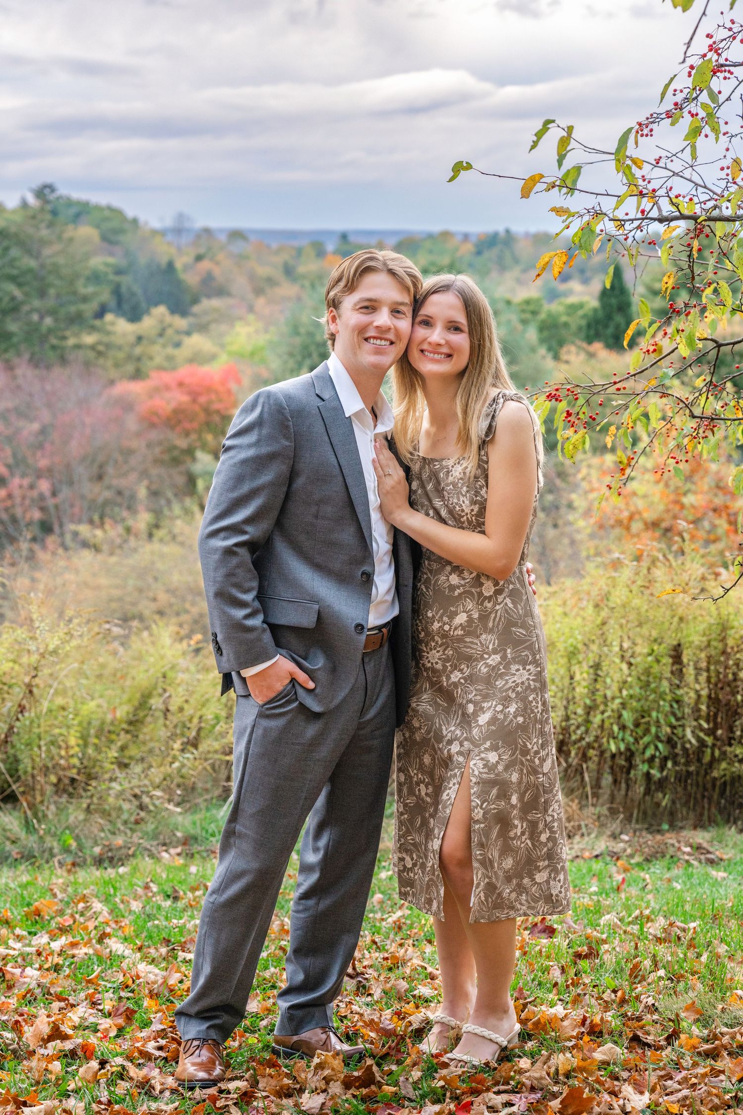 A couple poses together on an autumn hillside surrounded by colorful fall foliage and trees.
