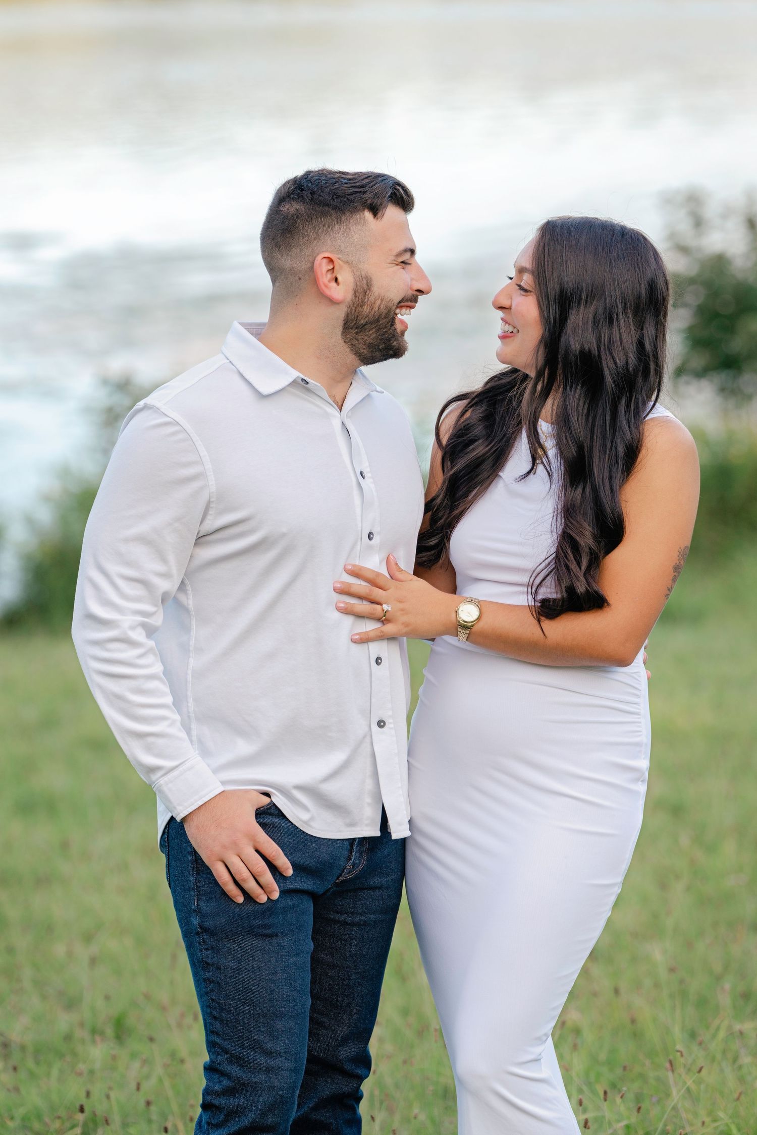A smiling couple in matching white outfits share a romantic moment outdoors near a scenic waterfront.