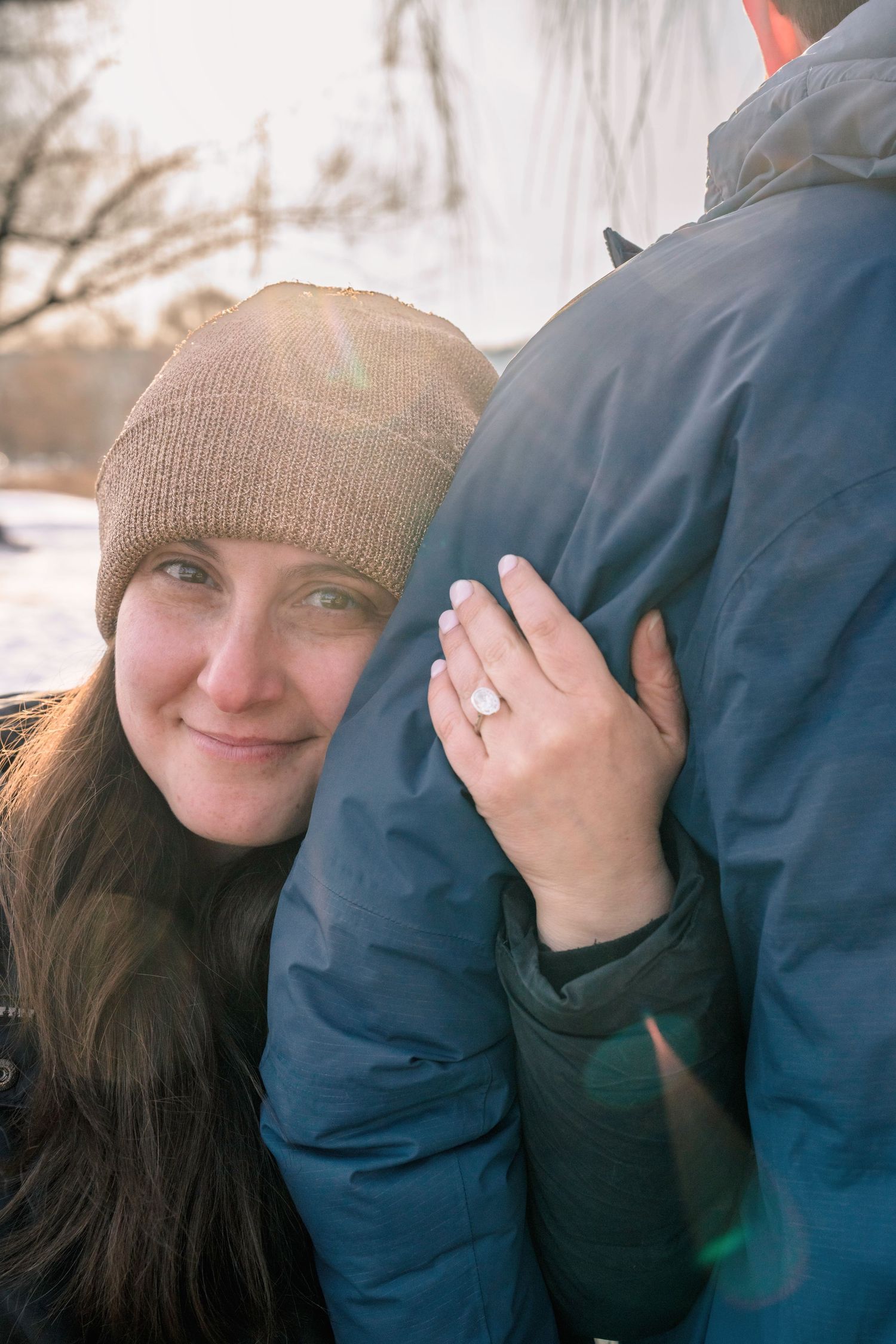 Woman in tan beanie hugs man outdoors, showing off engagement ring, with warm sunlight in winter setting.