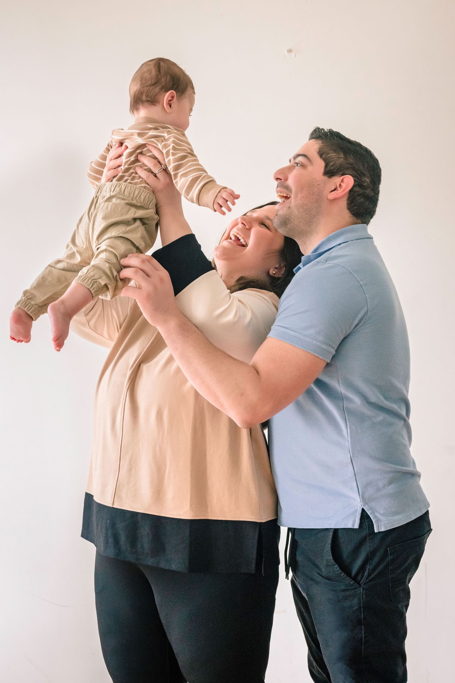 Joyful parents laugh together while lifting their baby up in a bright white room.