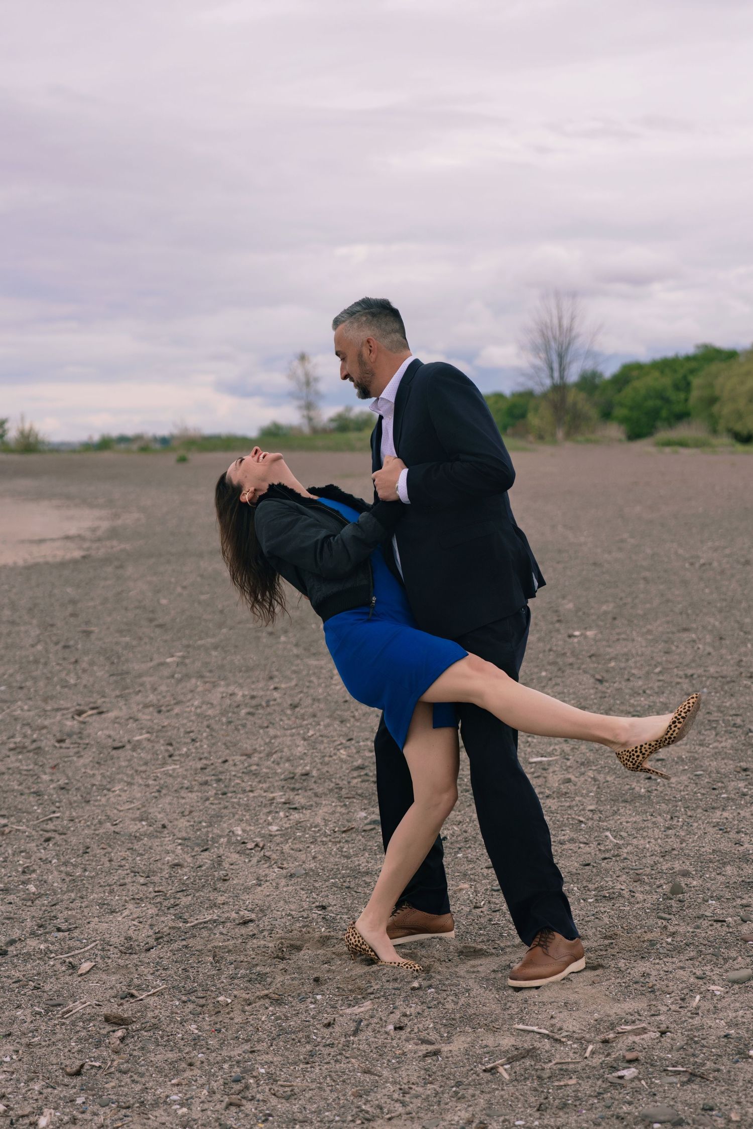 Playful dance sequence of a couple in formal attire on barren ground under cloudy skies.