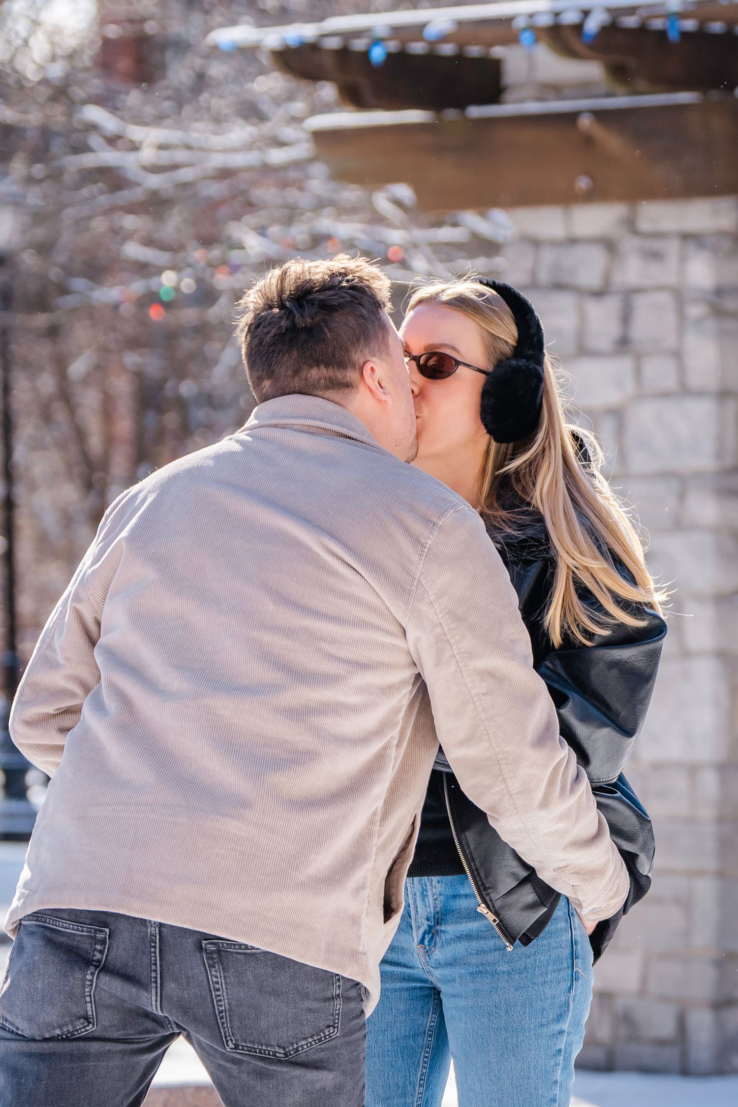 Romantic couple sharing a kiss outdoors in winter, woman wearing ear muffs and sunglasses near stone building.