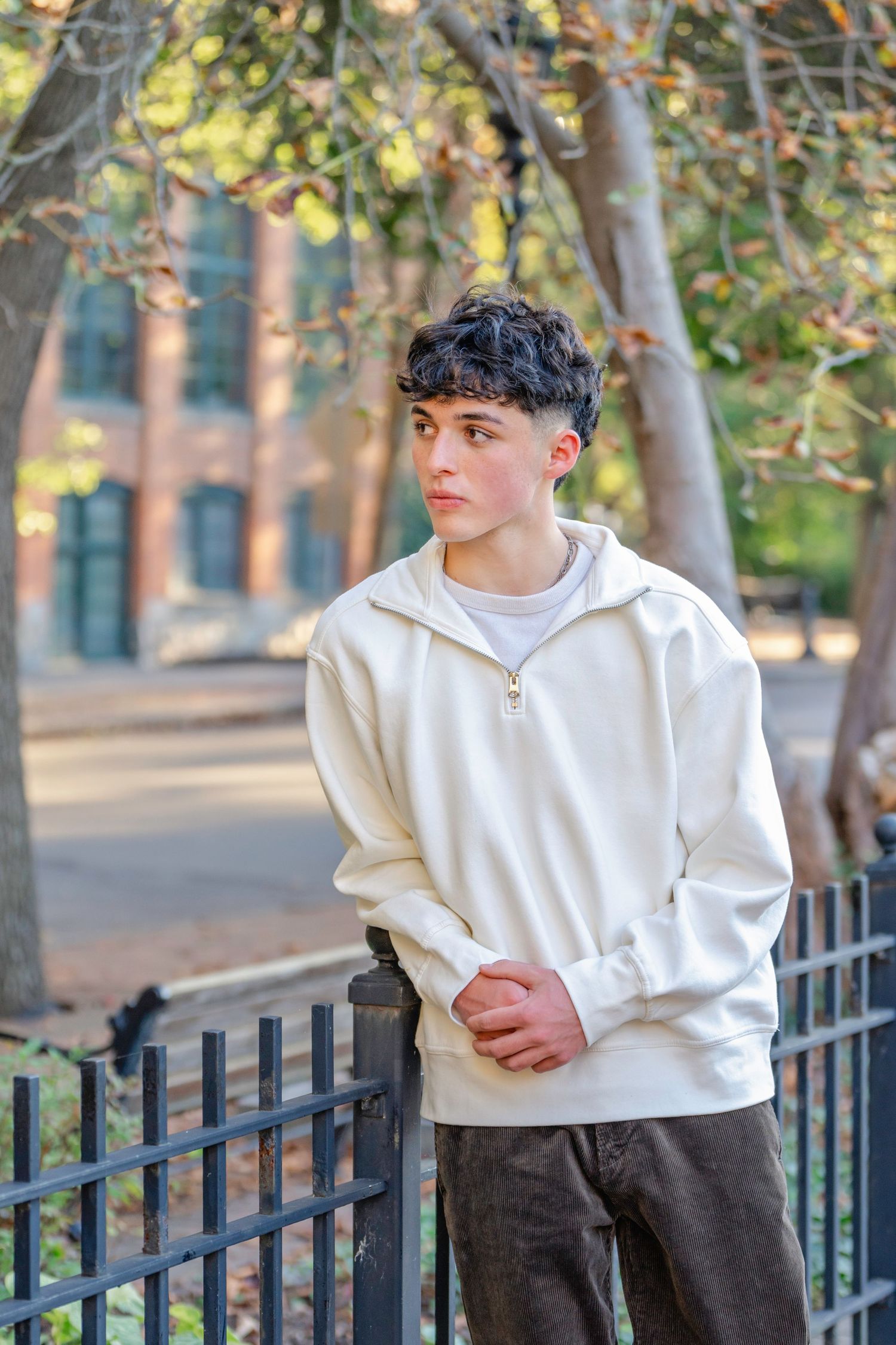 A person in a white hoodie and dark pants poses against a black iron fence with trees in the background.
