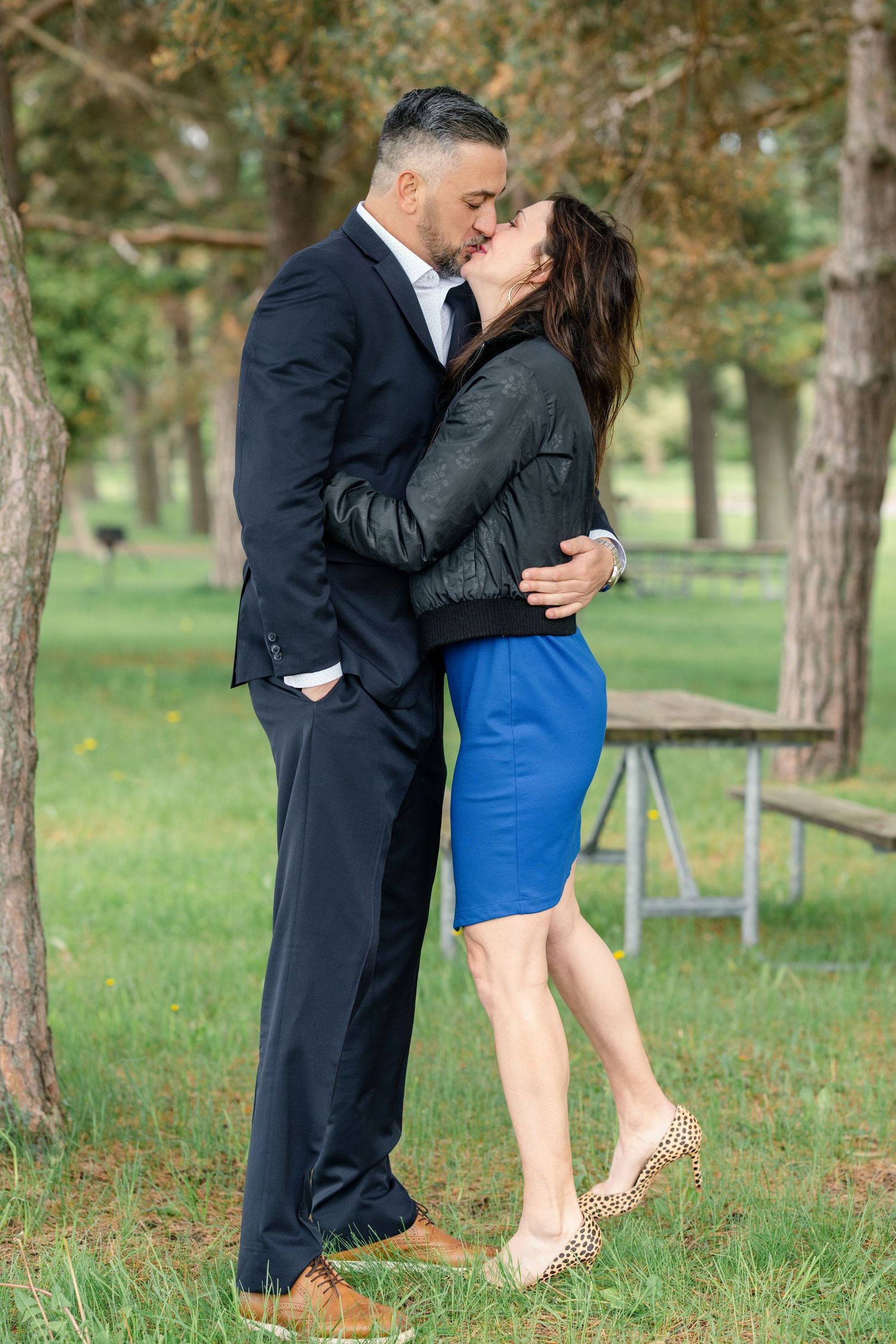A romantic couple shares tender kisses in a park setting with pine trees and green grass in the background.