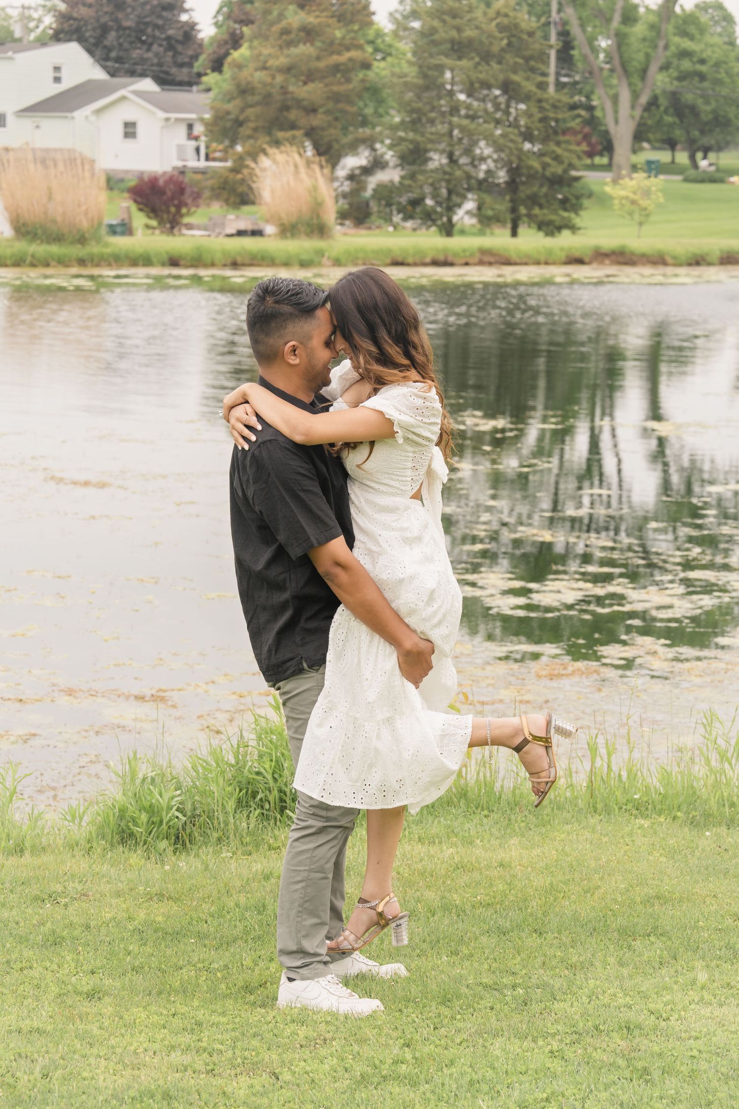 Playful couple dancing and embracing by a serene lake with lush greenery in the background.