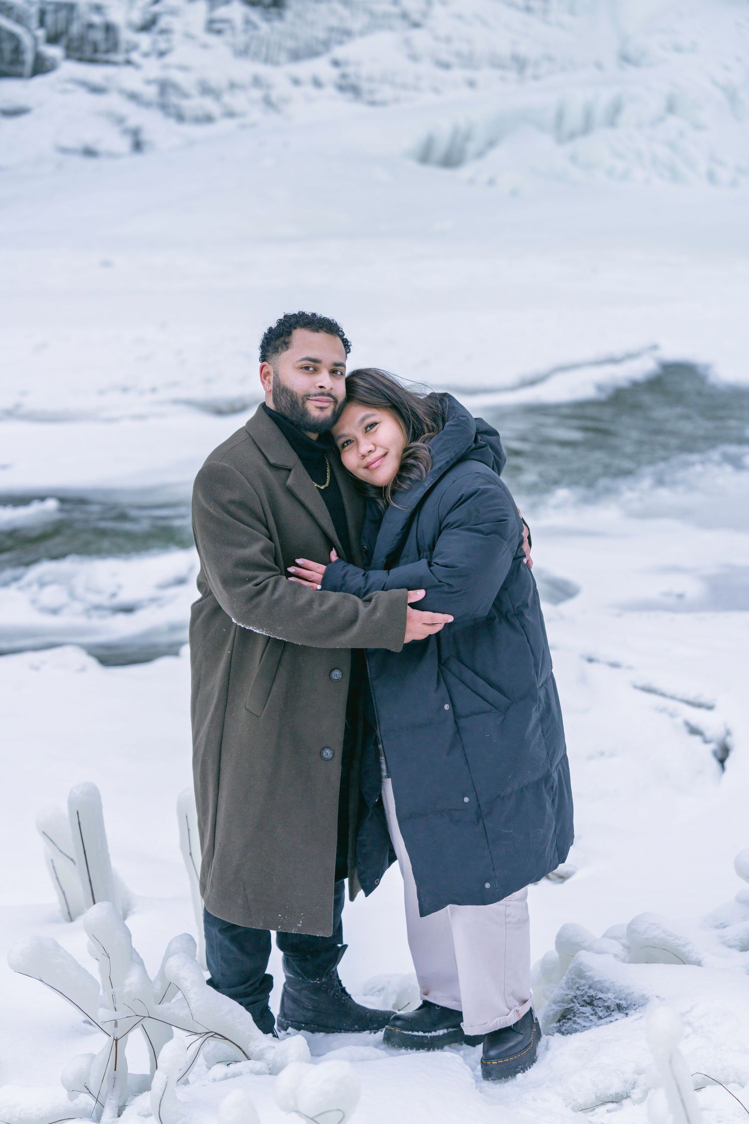 A couple embraces while sitting in a snowy winter landscape with ice formations in the background.