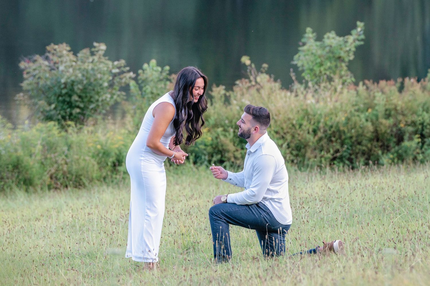 Couple in white outfits takes engagement photos in a scenic grassy meadow during golden hour.