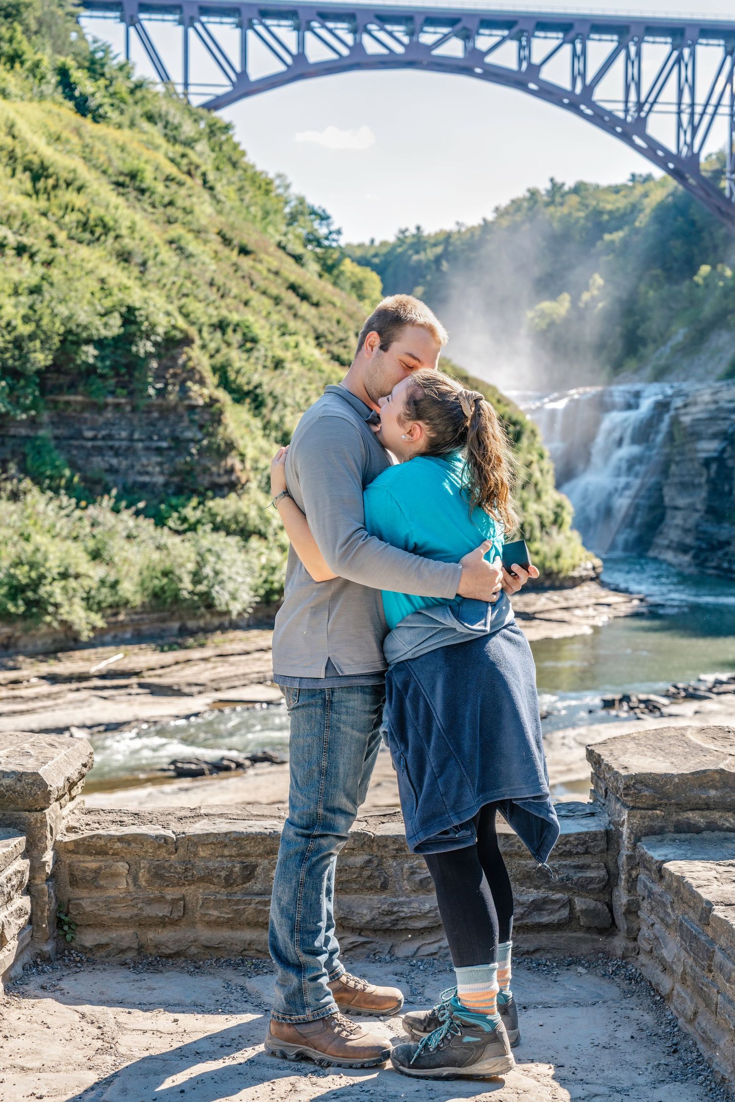 A couple embraces near a scenic waterfall and bridge overlook on a sunny afternoon.