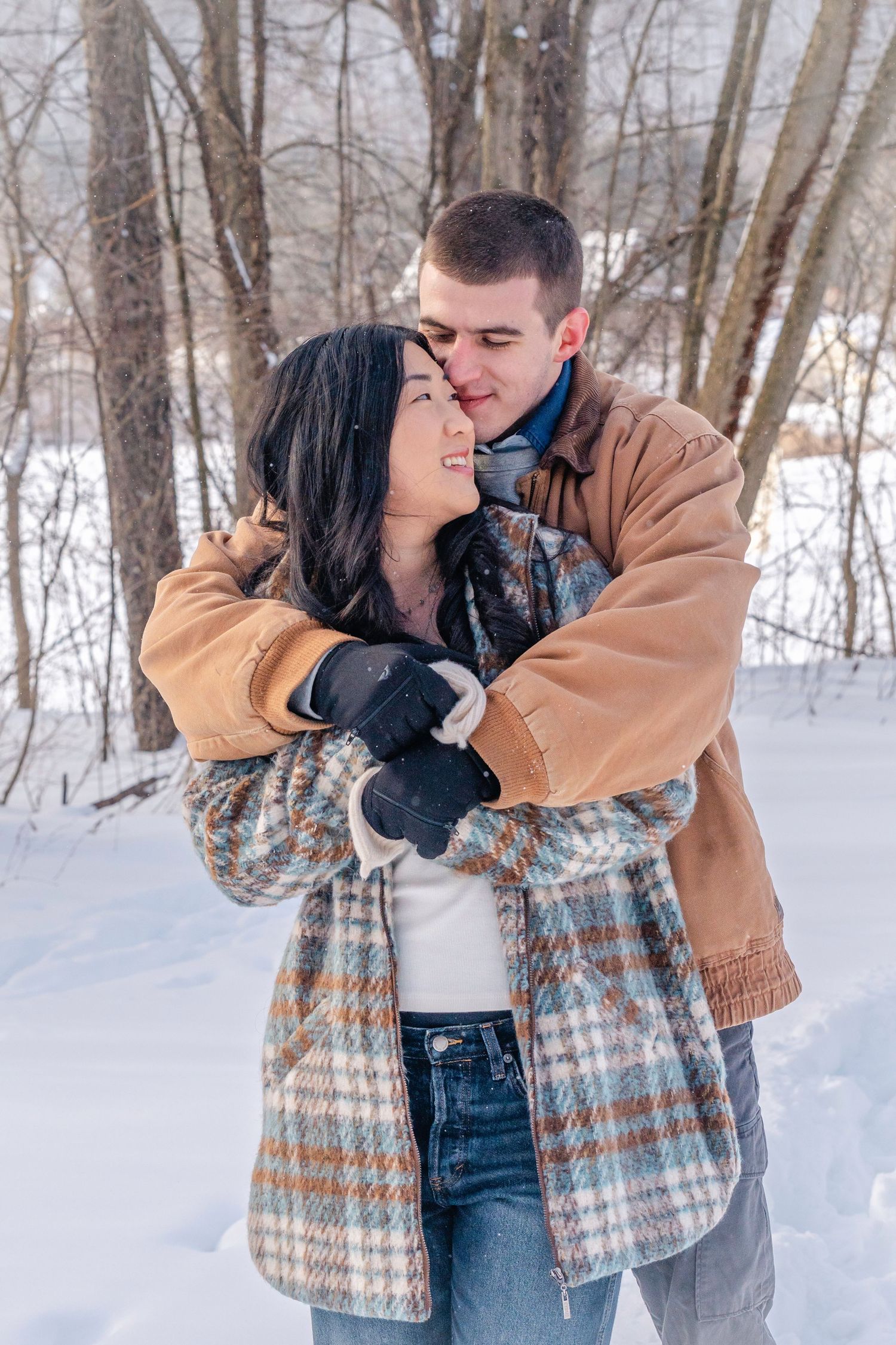 Couple embracing in snowy winter forest, wearing warm coats and gloves, sharing a romantic moment outdoors.