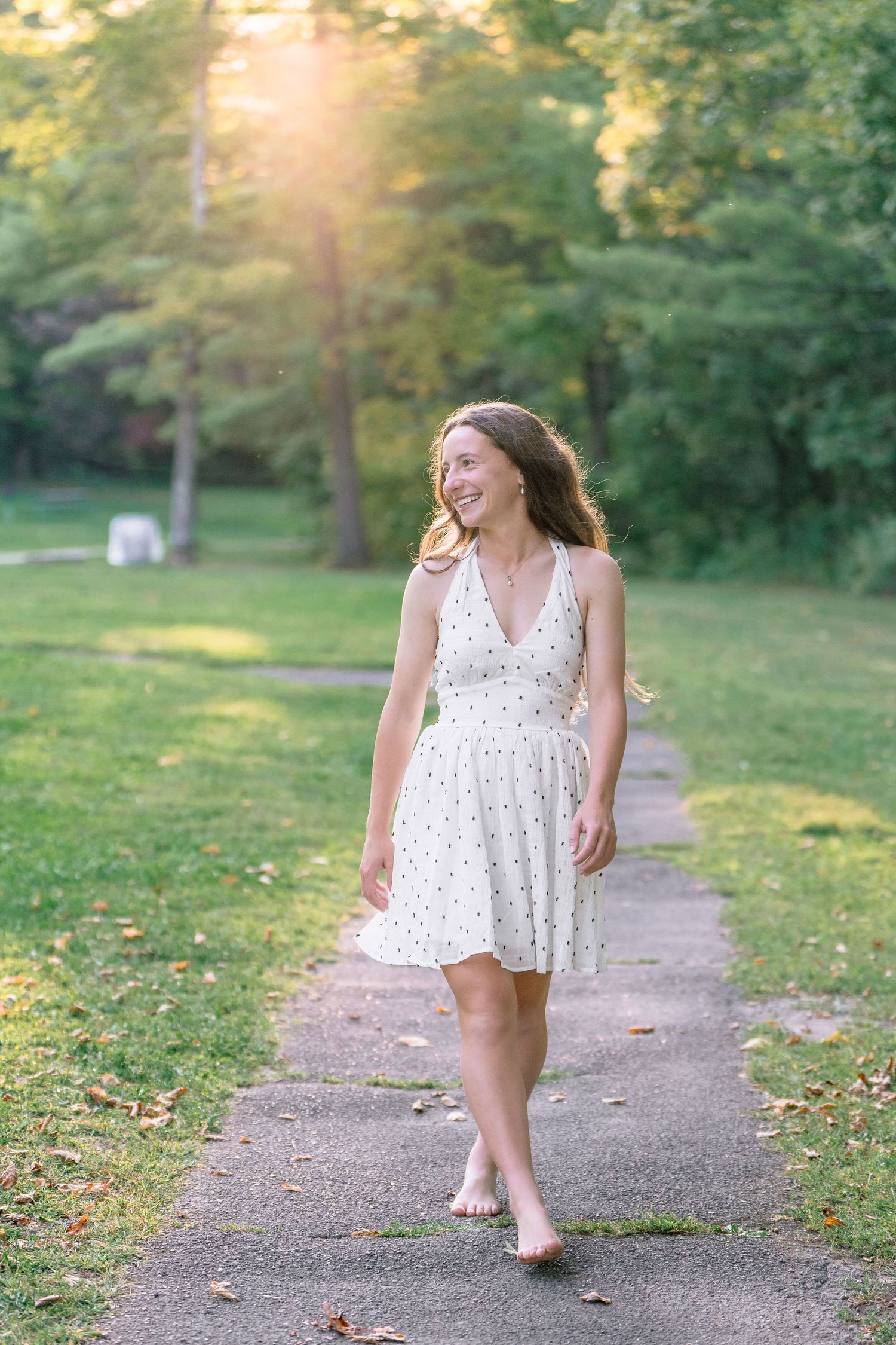 A high school senior in a flowing white polka dot dress twirls playfully on a sunlit garden path during golden hour.