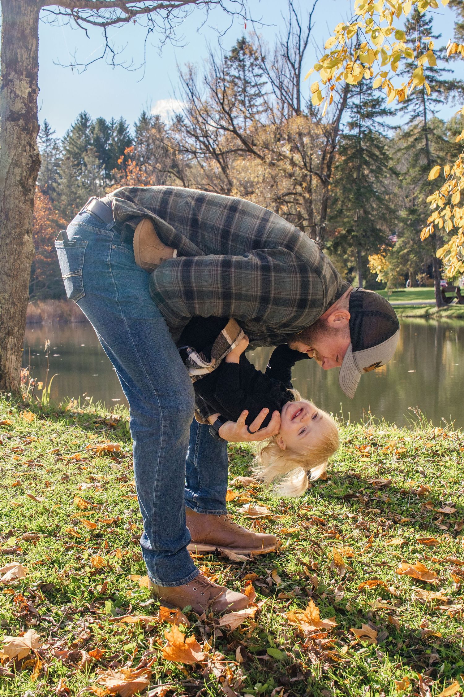 A person in plaid shirt and jeans bends down to pick up autumn leaves by a serene lake on a sunny fall day.