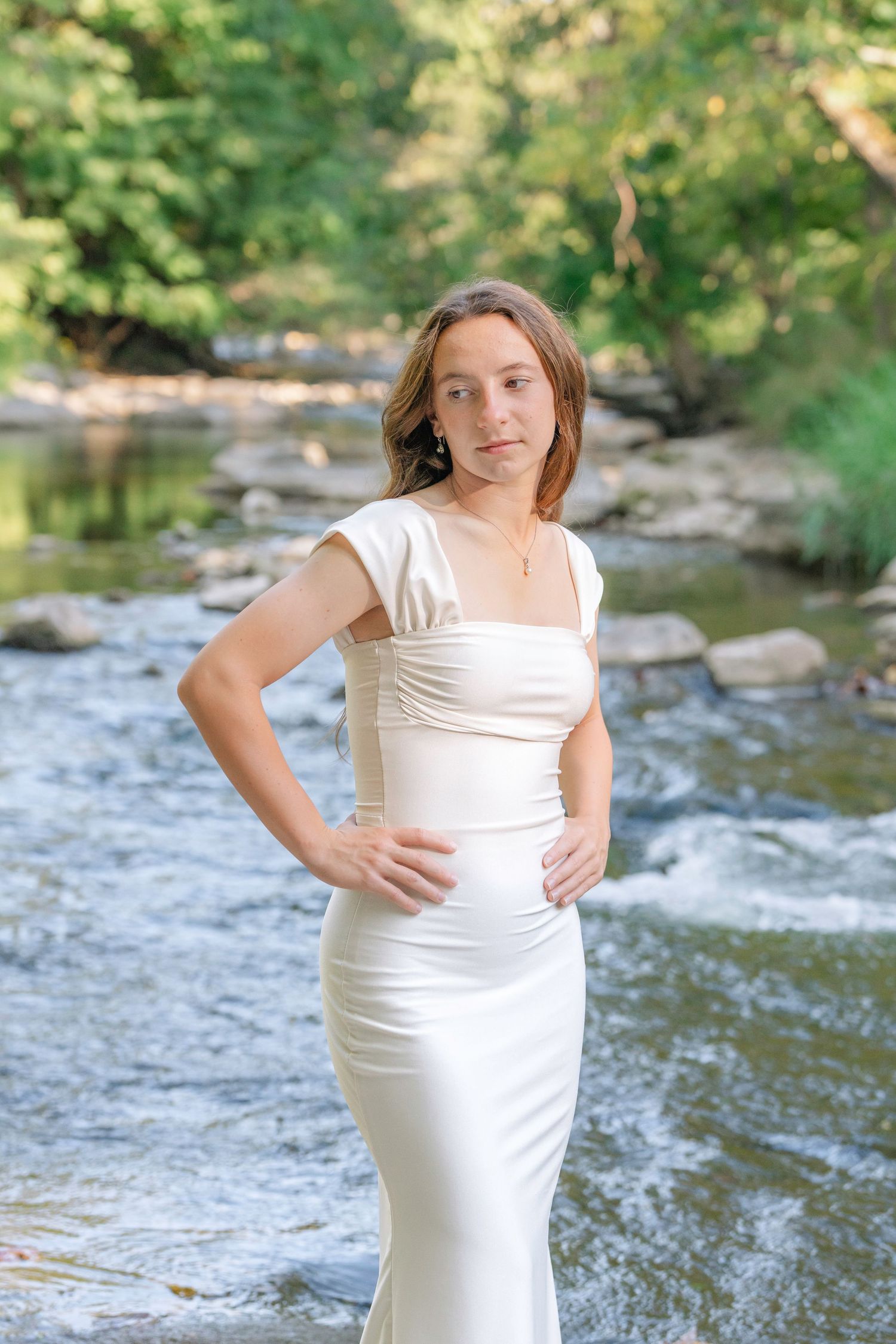 A person in a white dress poses by a scenic creek with rocks and trees in the background.