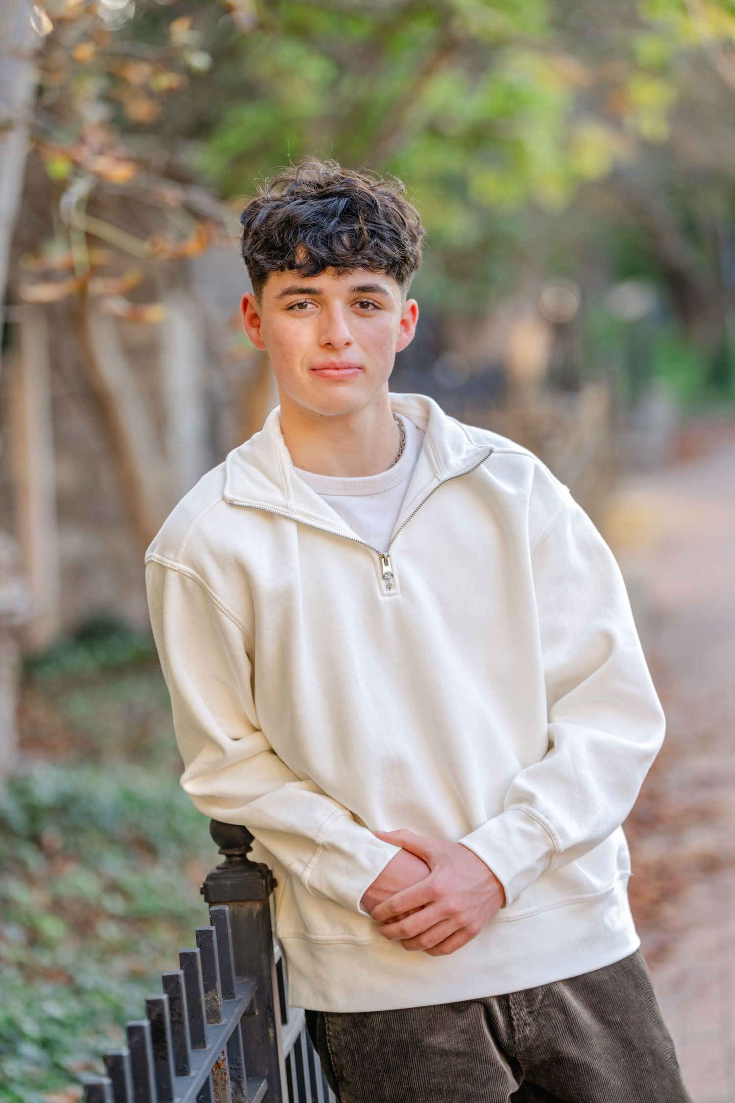 Person in white half-zip sweater and dark pants poses outdoors on wooded path in autumn.