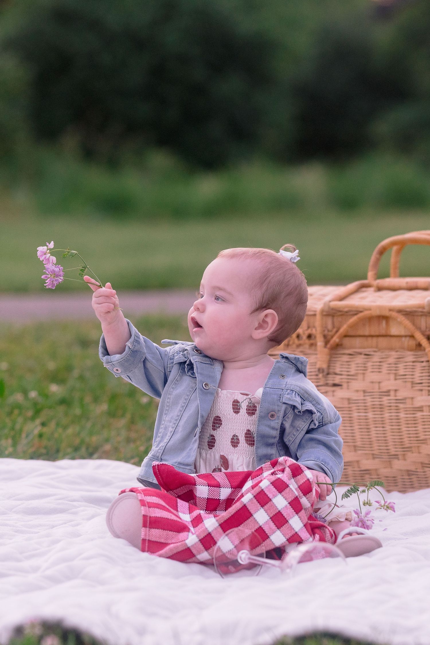 Baby in denim jacket holds small flowers while sitting on white blanket with picnic basket.