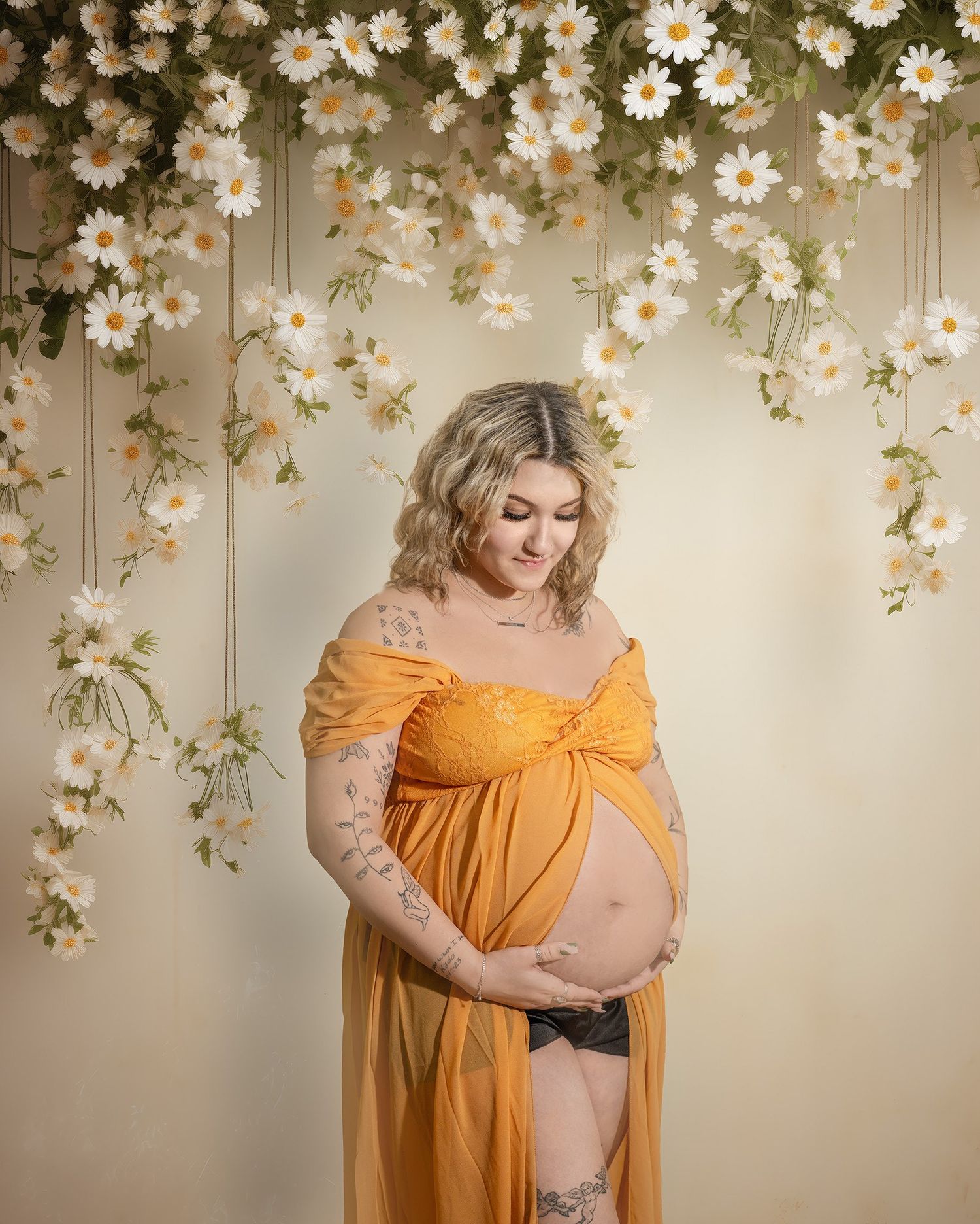 A maternity photo with flowing yellow dress against a backdrop of cascading white daisies in a dreamy indoor setting.