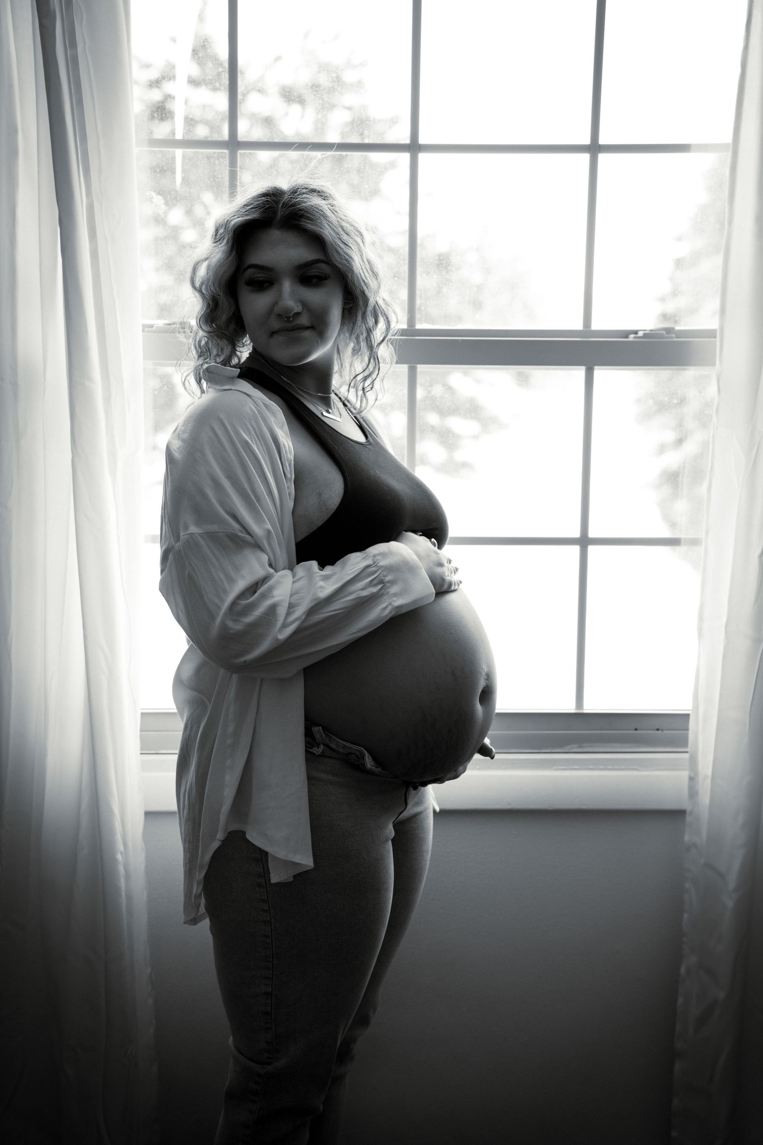 Black and white maternity photo by a window showing an expectant mother in a white button-down shirt cradling her belly.