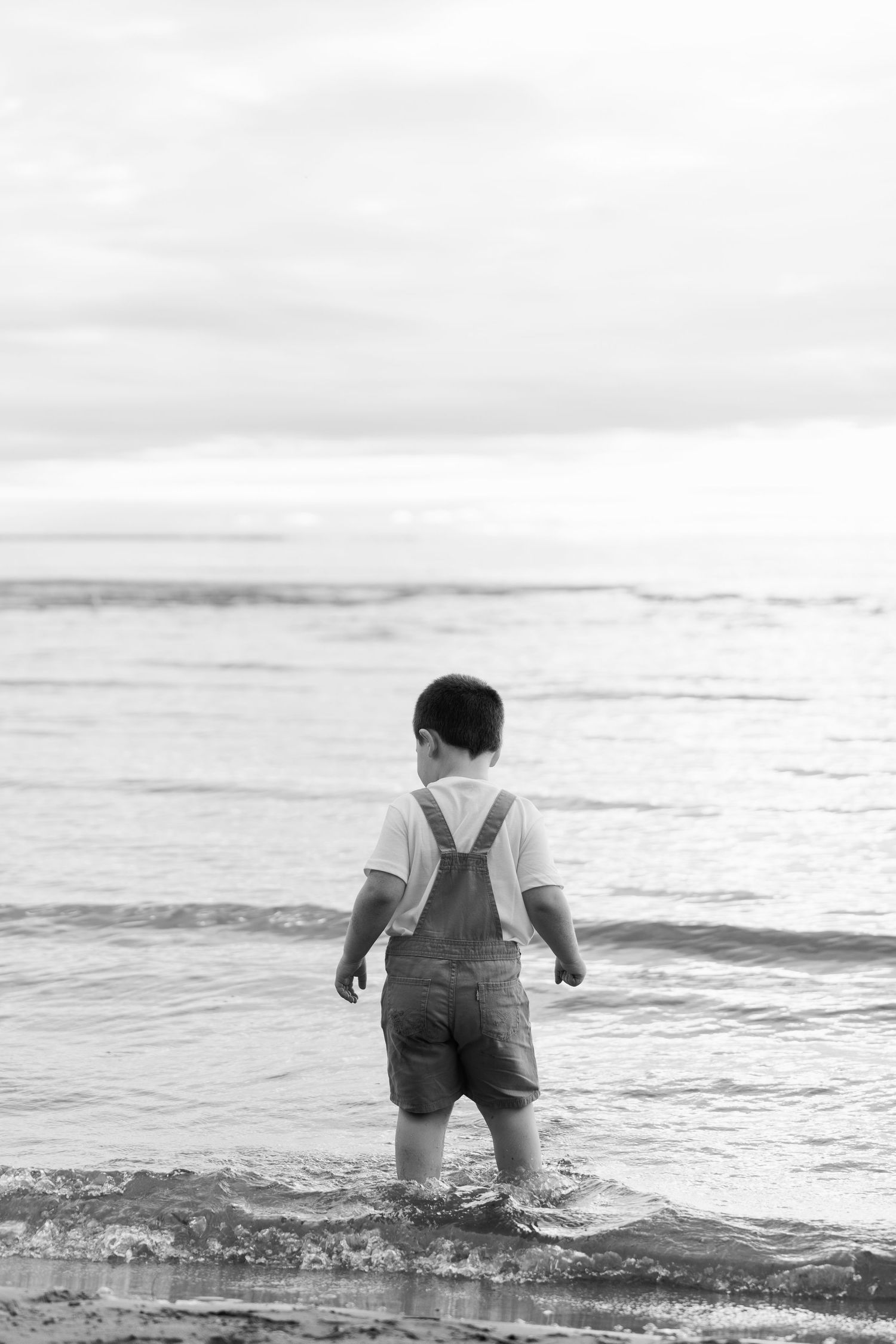 A small child wades into the ocean waves in black and white, creating a nostalgic beach scene.