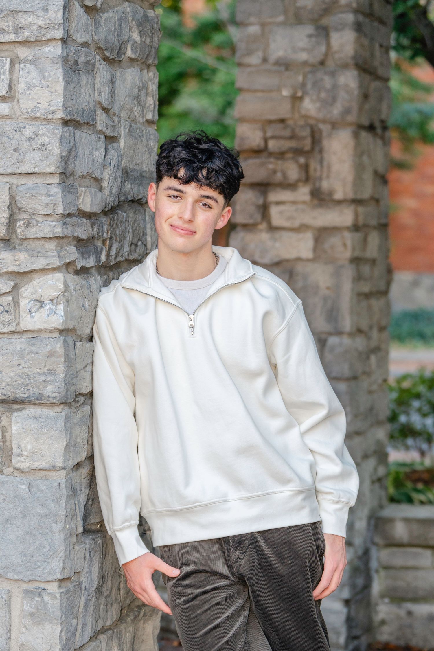 A casual portrait against a stone wall backdrop features someone in a white quarter-zip sweatshirt and dark pants.