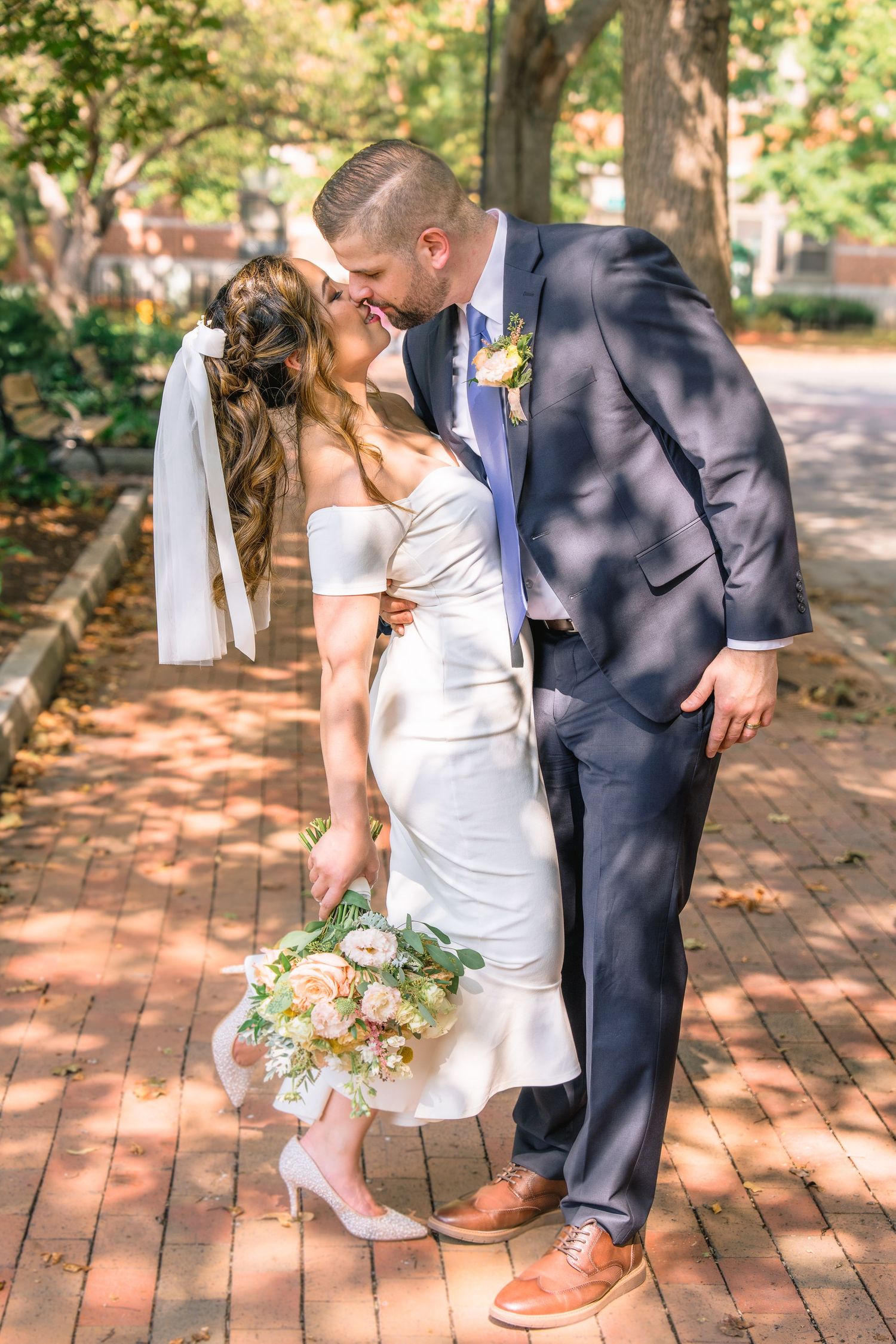 A romantic wedding couple embraces on a brick pathway surrounded by dappled sunlight and greenery.