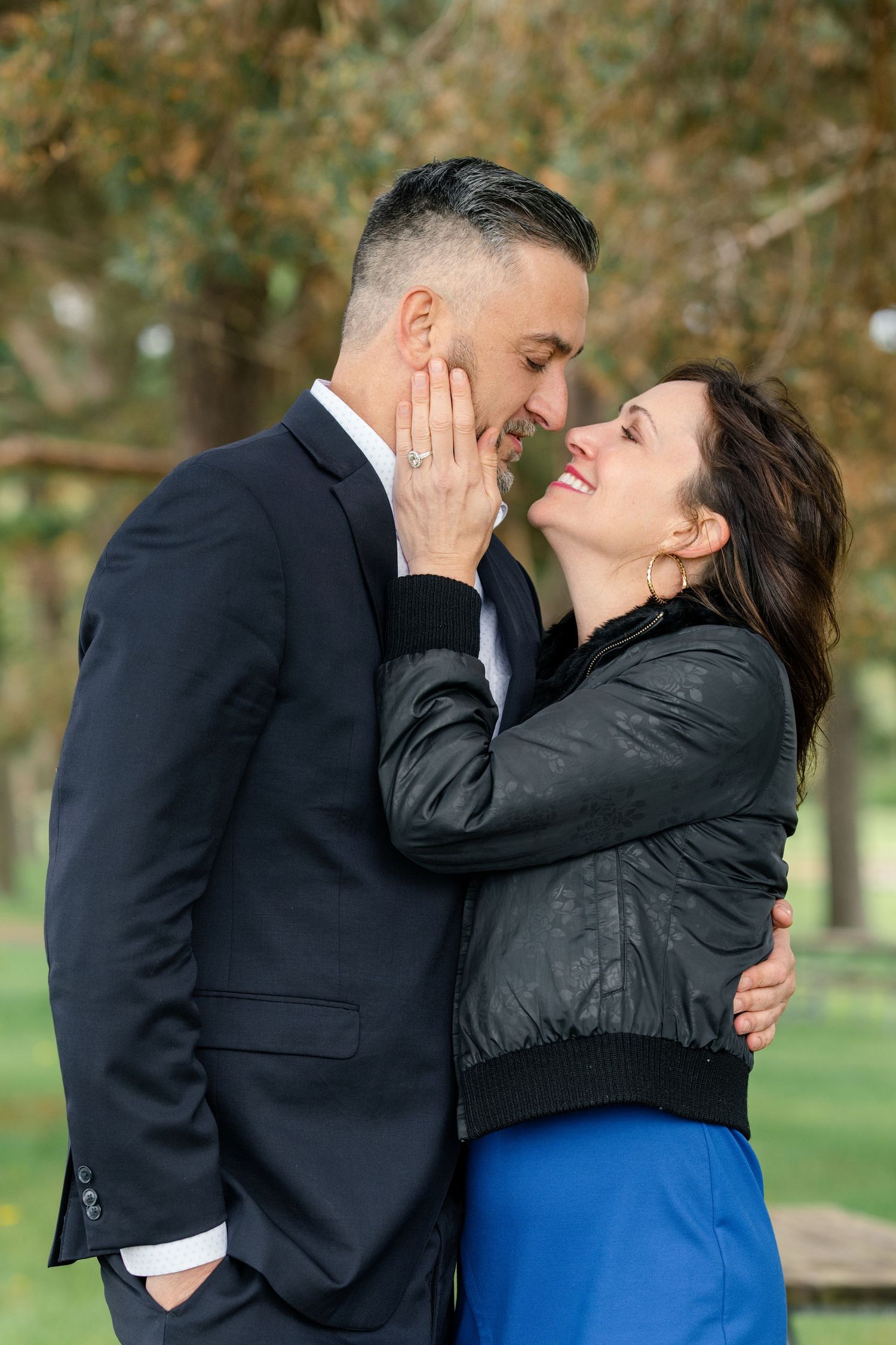 A couple shares an intimate moment in a park setting, with her in a black jacket and him in formal attire.