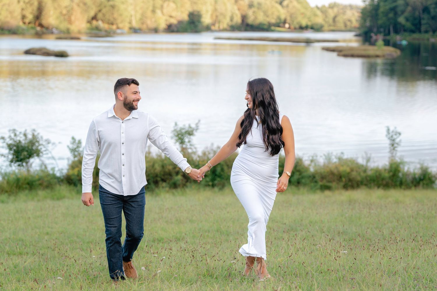 A couple embraces on grassy terrain with a serene lake and trees in the background.